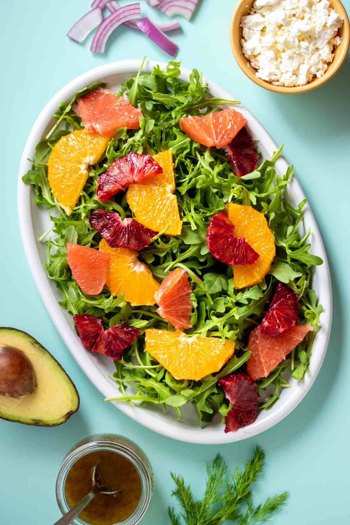 A salad with citrus pieces on a white platter with salad ingredients around it on a turquoise background.