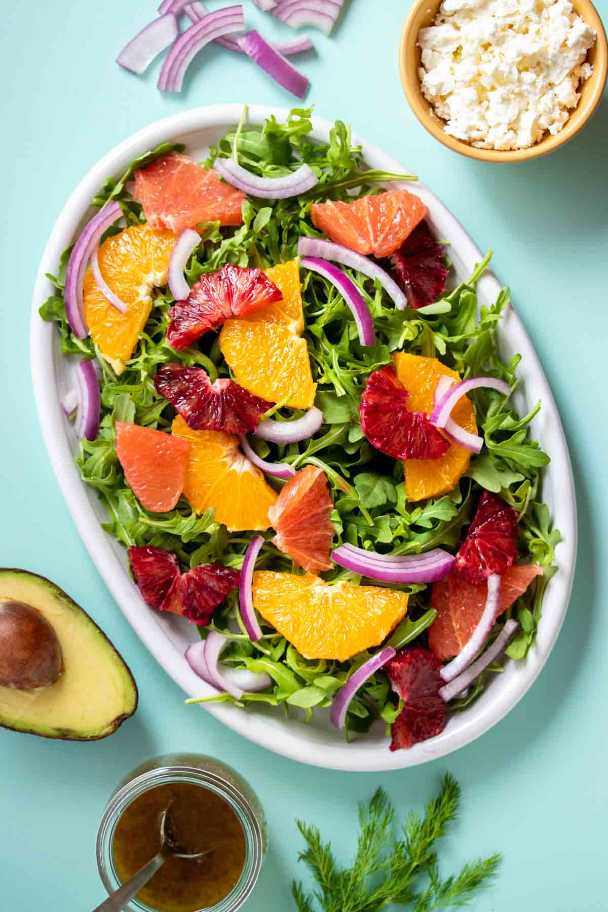 A white platter on a turquoise background with arugula, citrus slices and red onion on top.