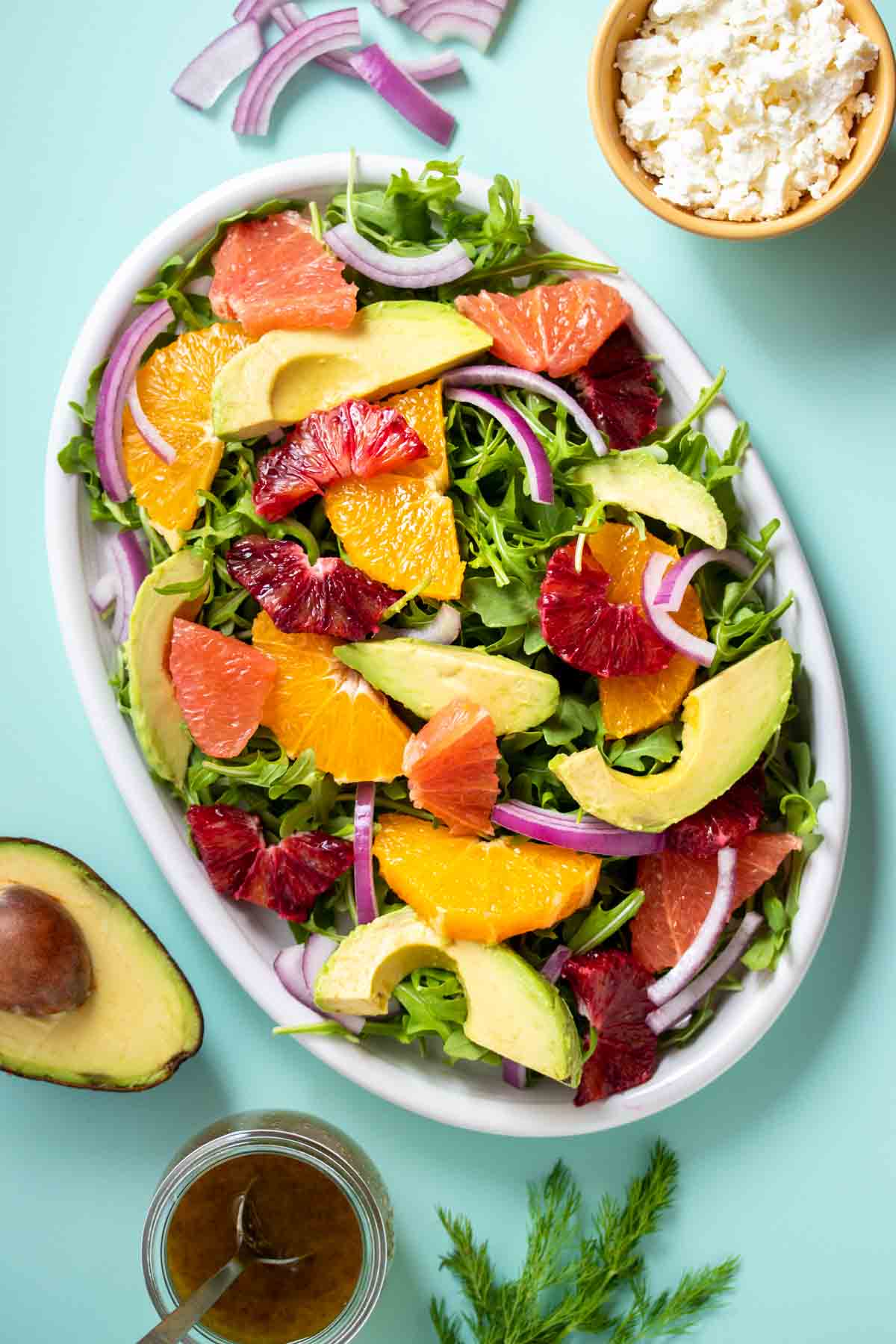 A turquoise background with a citrus salad with onion and avocado on it and other ingredients around the plate.