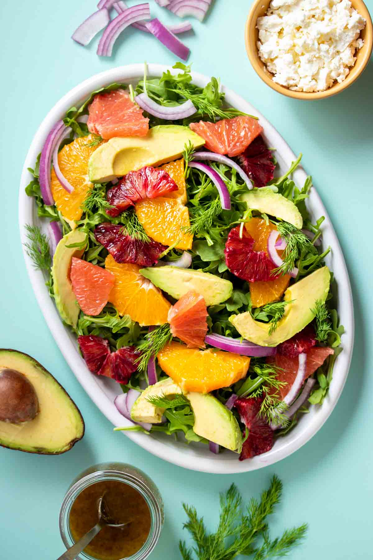 White plate with a citrus and avocado salad on it sitting on a turquoise background with other ingredients around it.