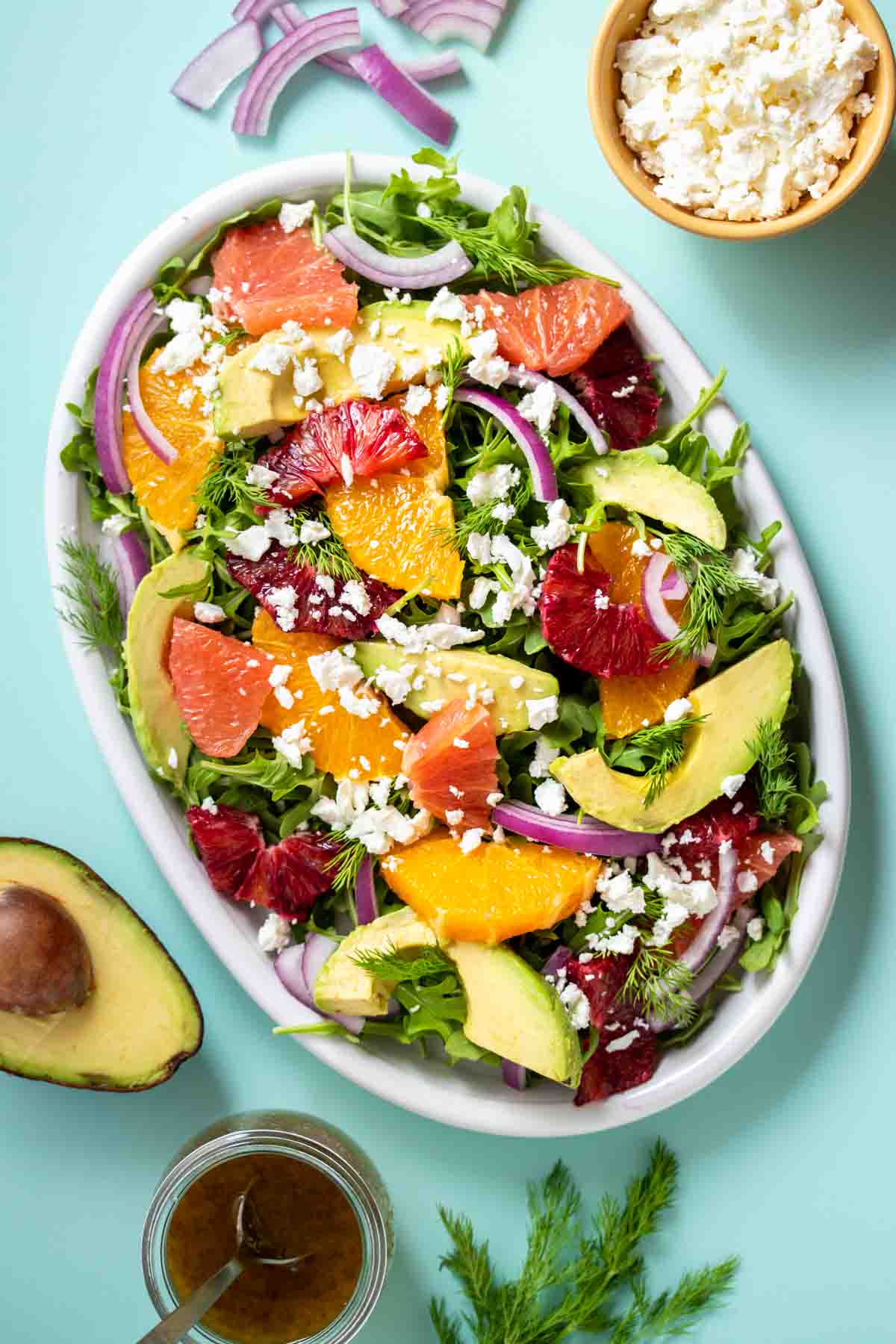 A citrus salad with avocado, red onion and feta on it sitting on a platter on a turquoise background with ingredients around it.