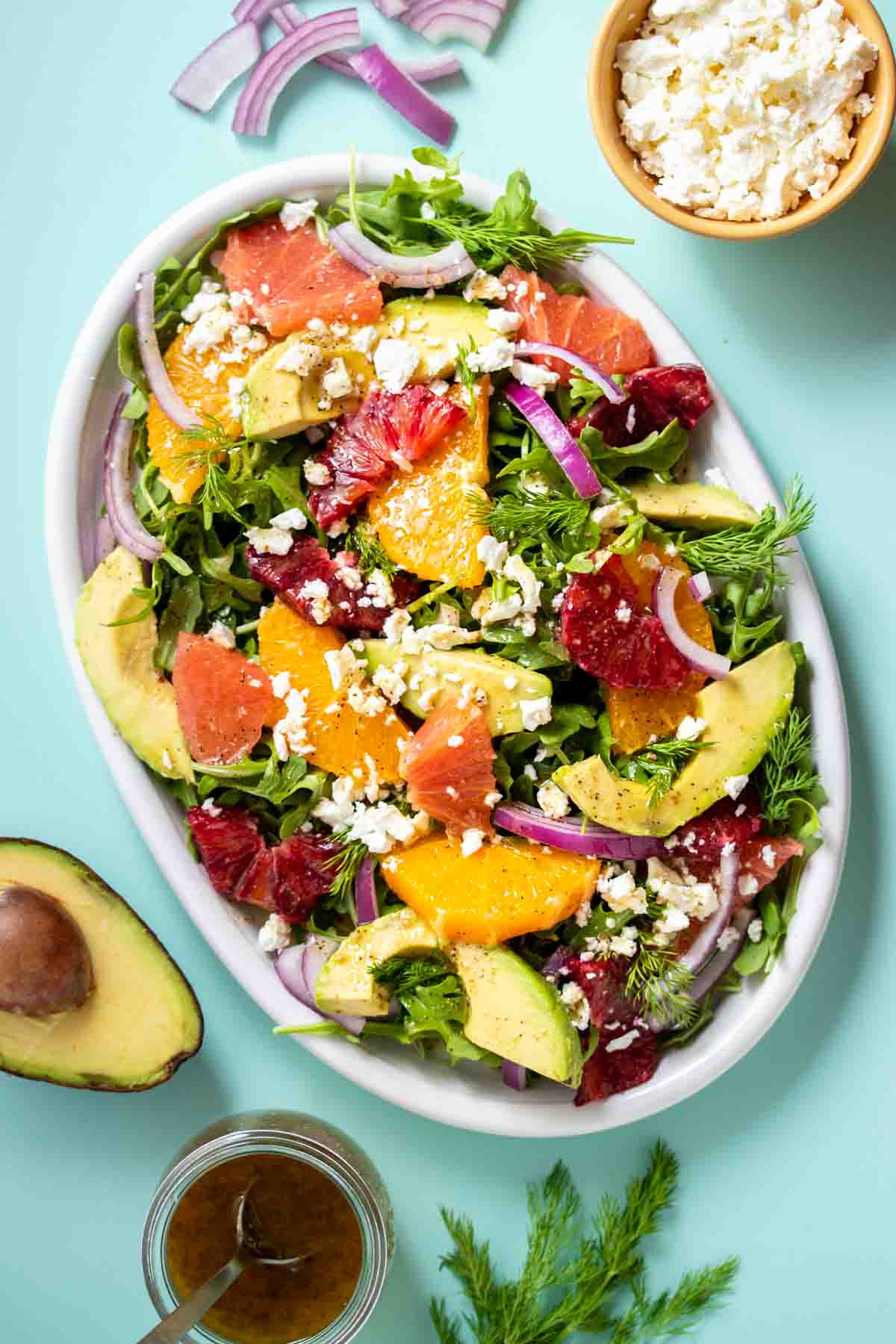 A citrus, feta, avocado and red onion salad with dressing on a white platter on a turquoise background with ingredients around it.