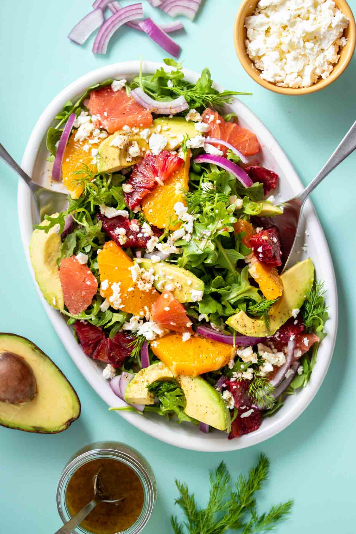 Silver spoons about to serve a salad with arugula, citrus and feta on a white platter on a turquoise background.