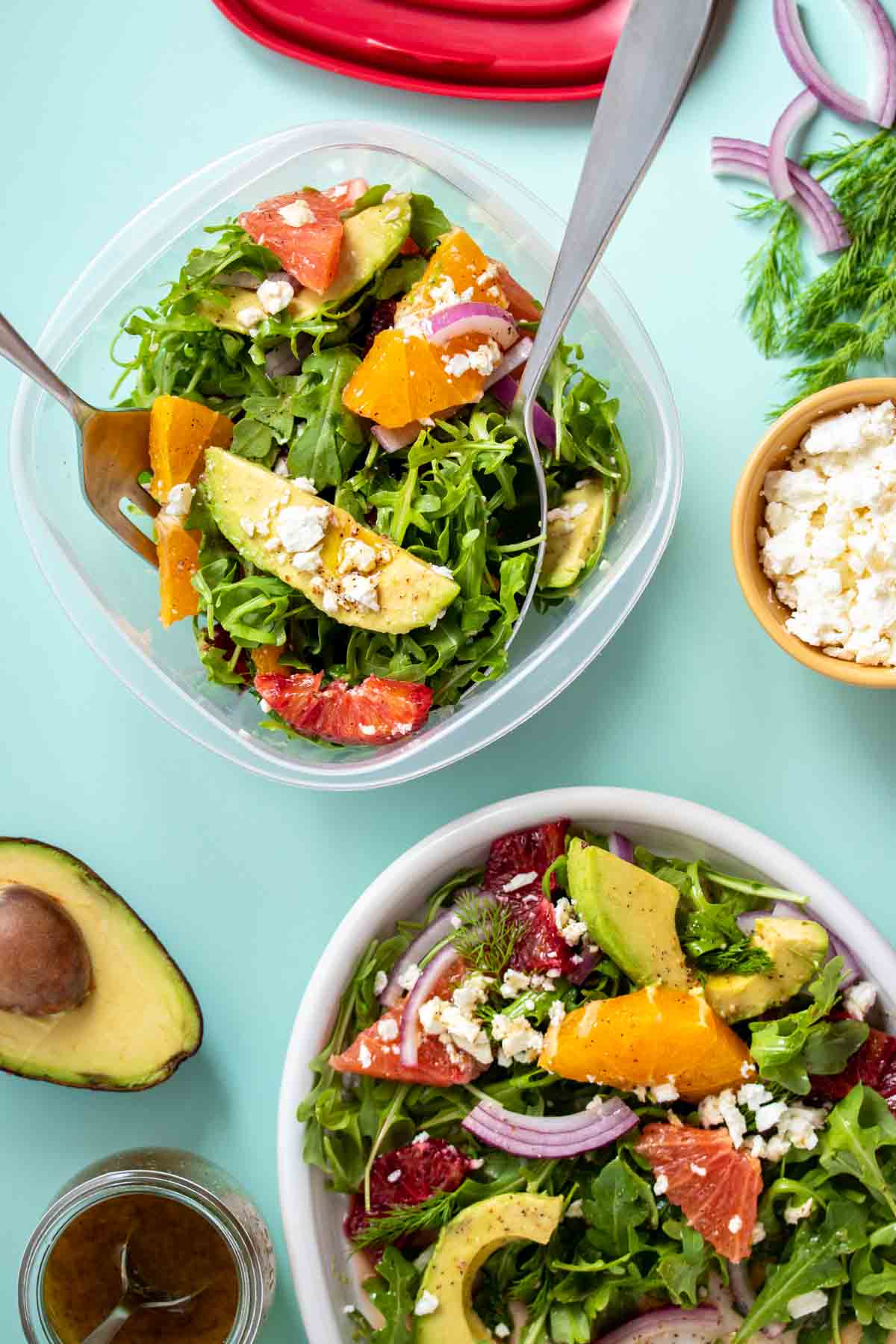 A citrus avocado and feta salad being put into a plastic square container from a white platter on a turquoise background.