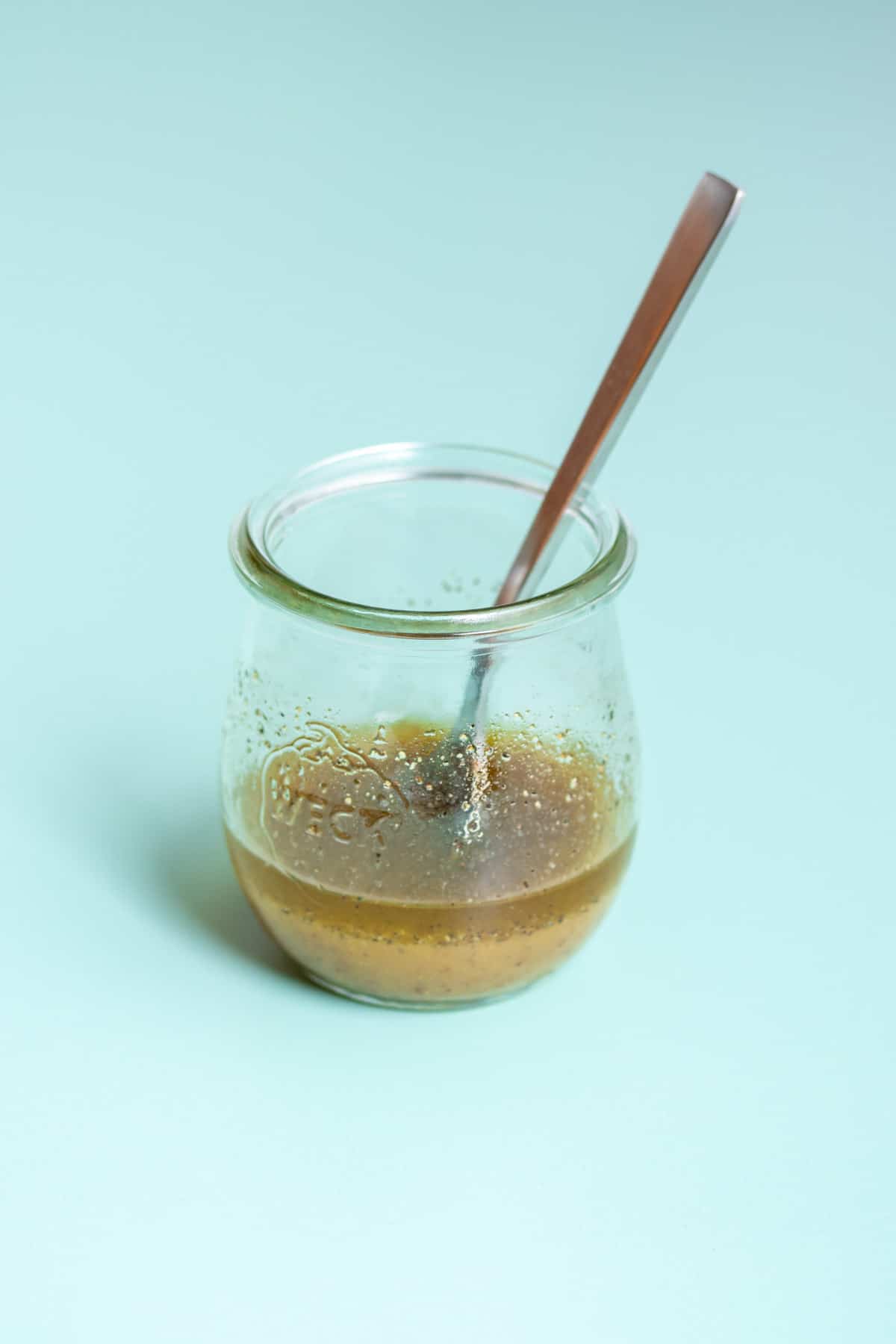 A glass jar with a light brown tan dressing in it and oil settled on the top on a turquoise background.