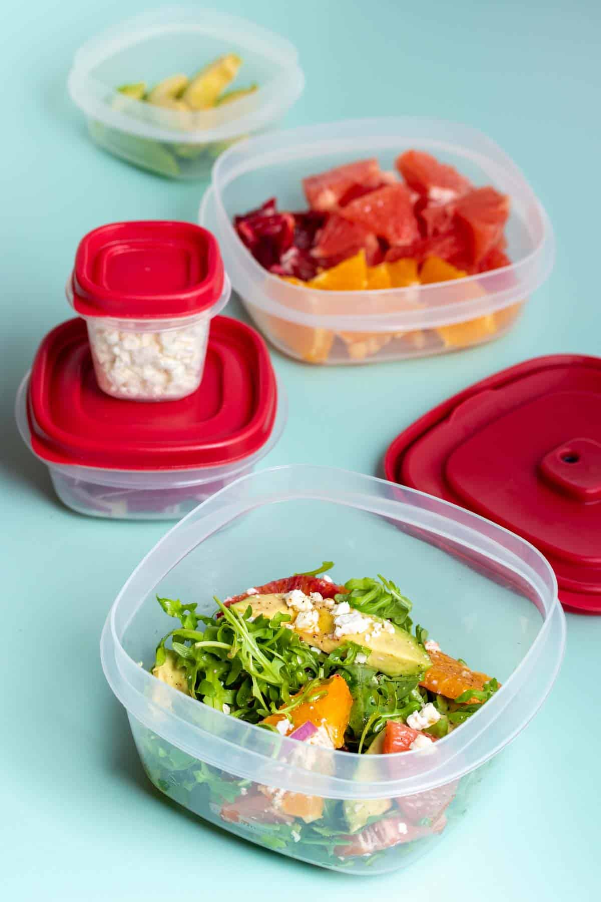 Plastic tuperware containers with red tops stacked and some open with ingredients for a citrus salad.