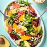 Spoons serving a citrus avocado and feta salad from a white platter with an avocado and bowl of feta next to it.