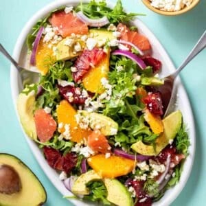 Spoons serving a citrus avocado and feta salad from a white platter with an avocado and bowl of feta next to it.