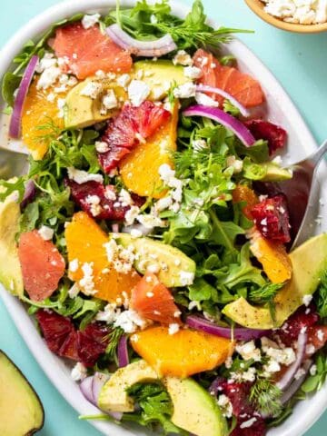 Spoons serving a citrus avocado and feta salad from a white platter with an avocado and bowl of feta next to it.