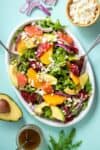 A citrus salad topped with avocado, feta, onion and dill next to half an avocado, jar of dressing, feta and onion.