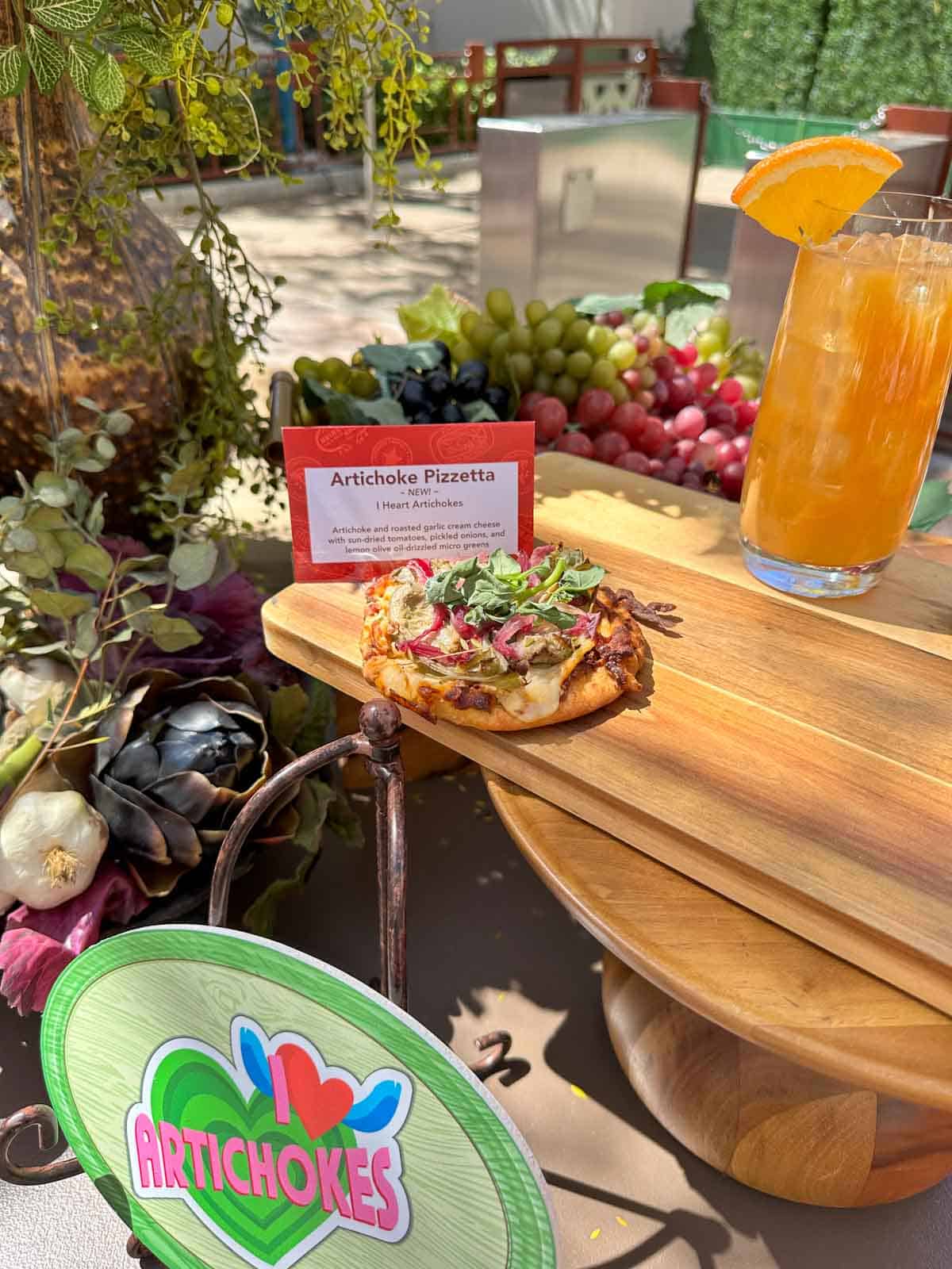 A round pizza style snack with artichokes, onion and basil on top on a wooden cutting board with a drink next to it.