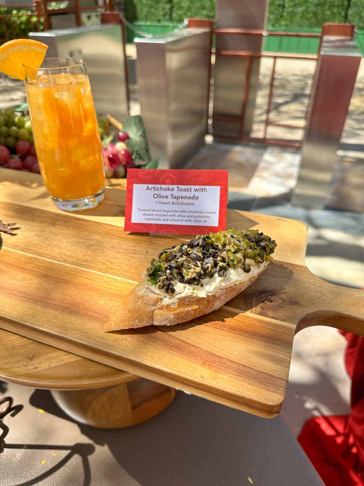A slice of bread with artichoke and olive chopped up on top sitting on a wooden cutting board outside.