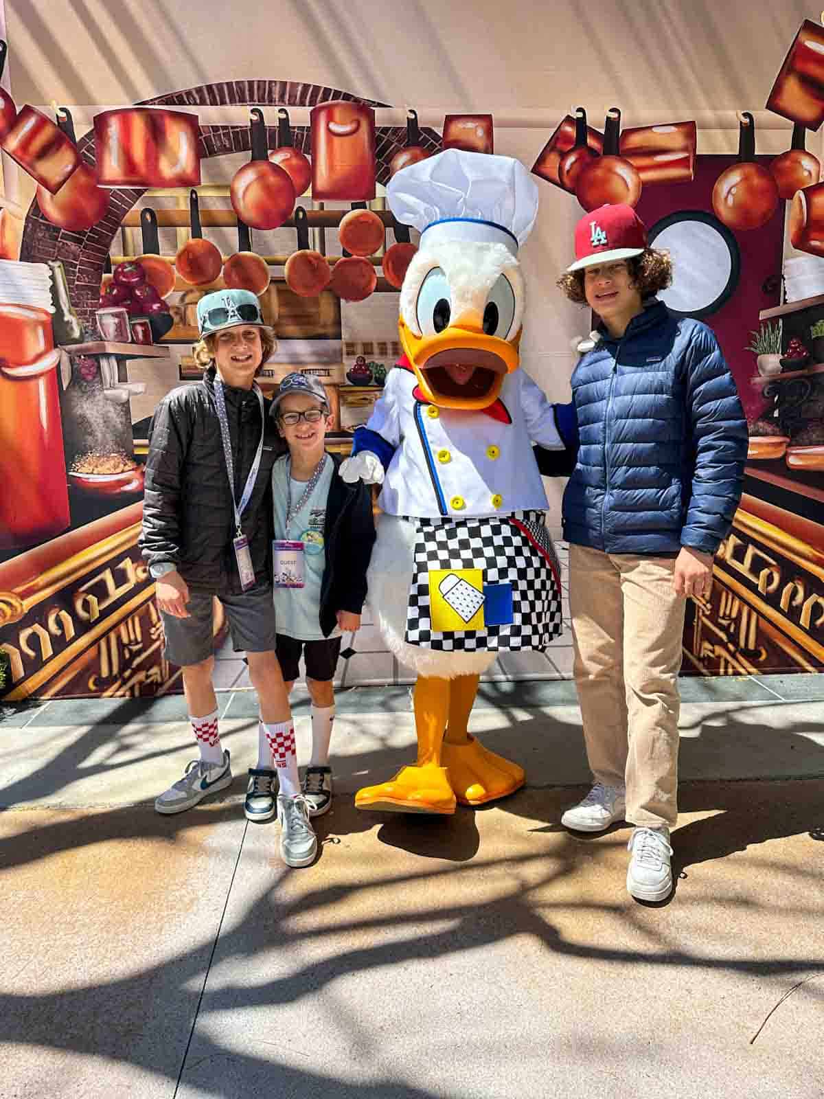 Donald Duck dressed as a chef with three boys smiling at the camera in front of a kitchen painting.