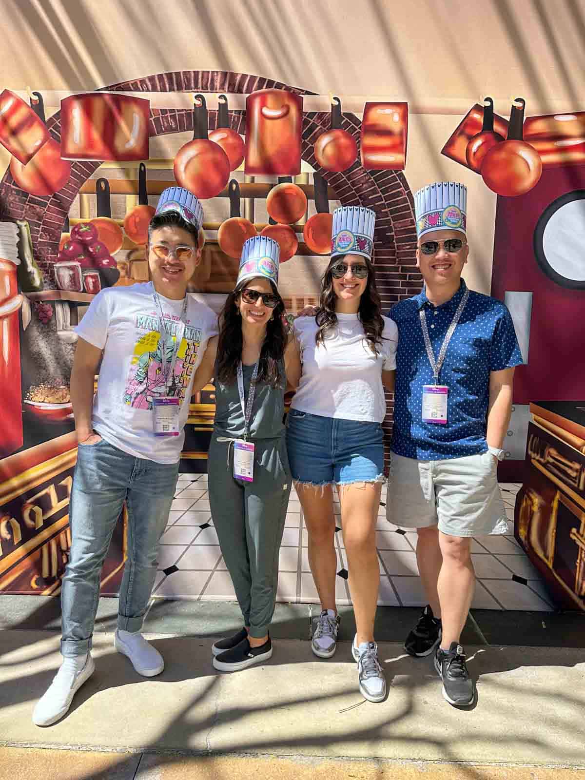 Four people with paper chef hats standing together smiling in front of a painting of a kitchen.