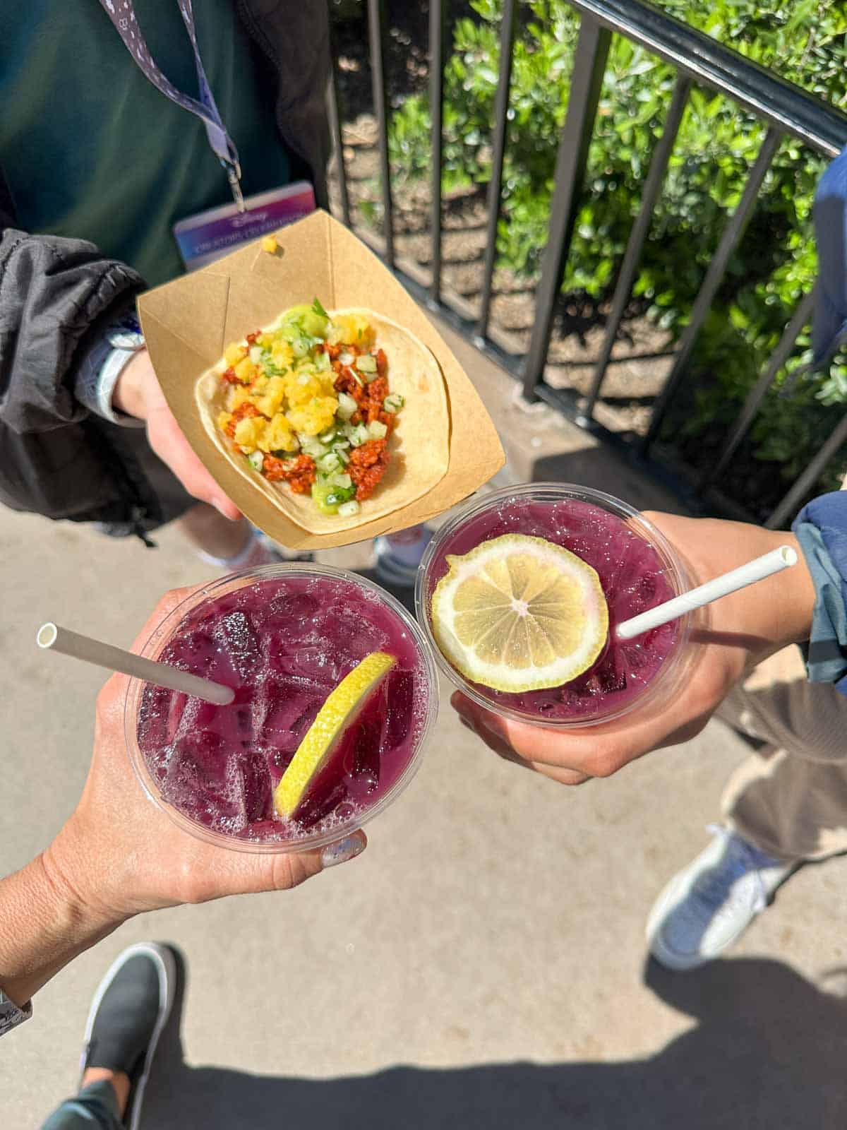 Hands doing cheers with purple drinks with a lemon on top and straws inside.