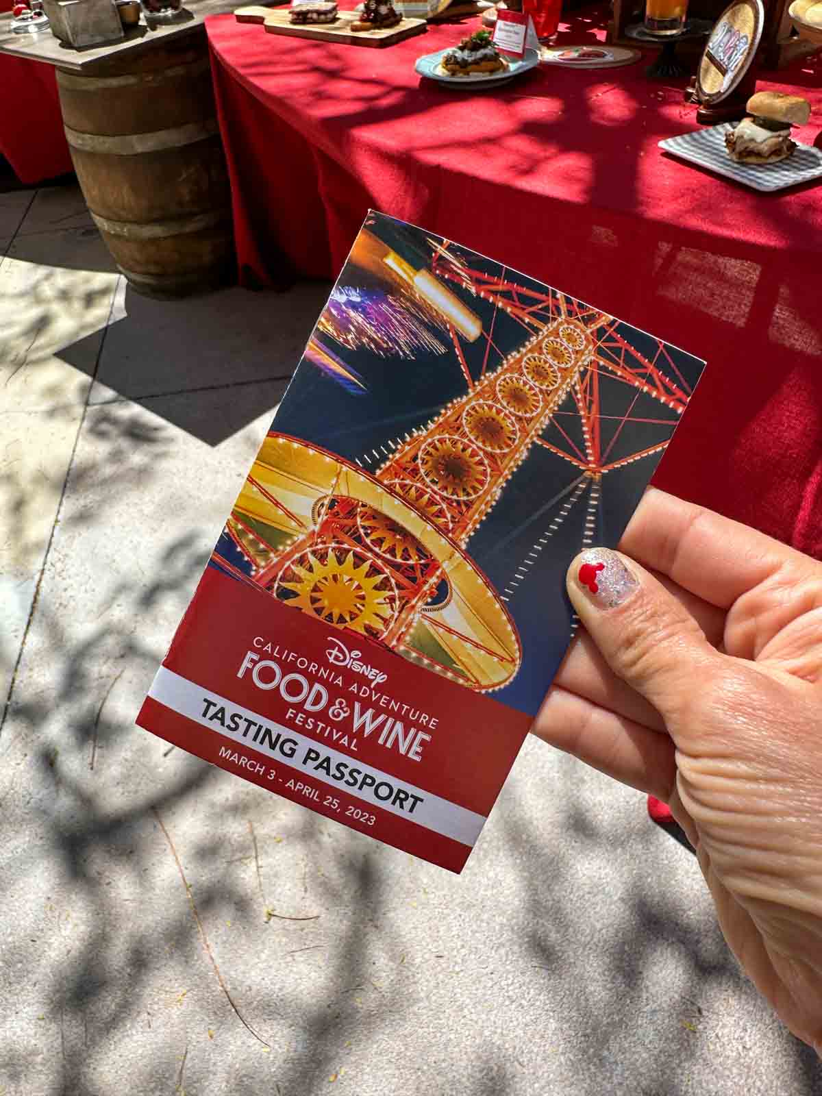 A hand holding a red tasting passport booklet for the Disney California Food and Wine festival with a photo of part of a ride on it.