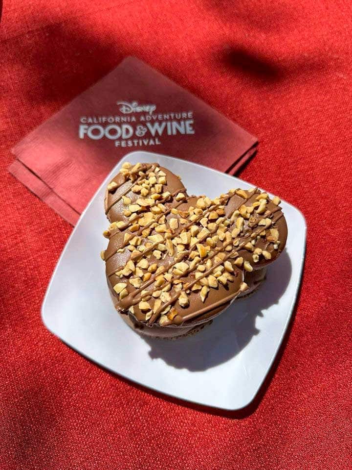 A mickey shaped chocolate dessert covered in nuts on top sitting on a white plate on a red tablecloth.