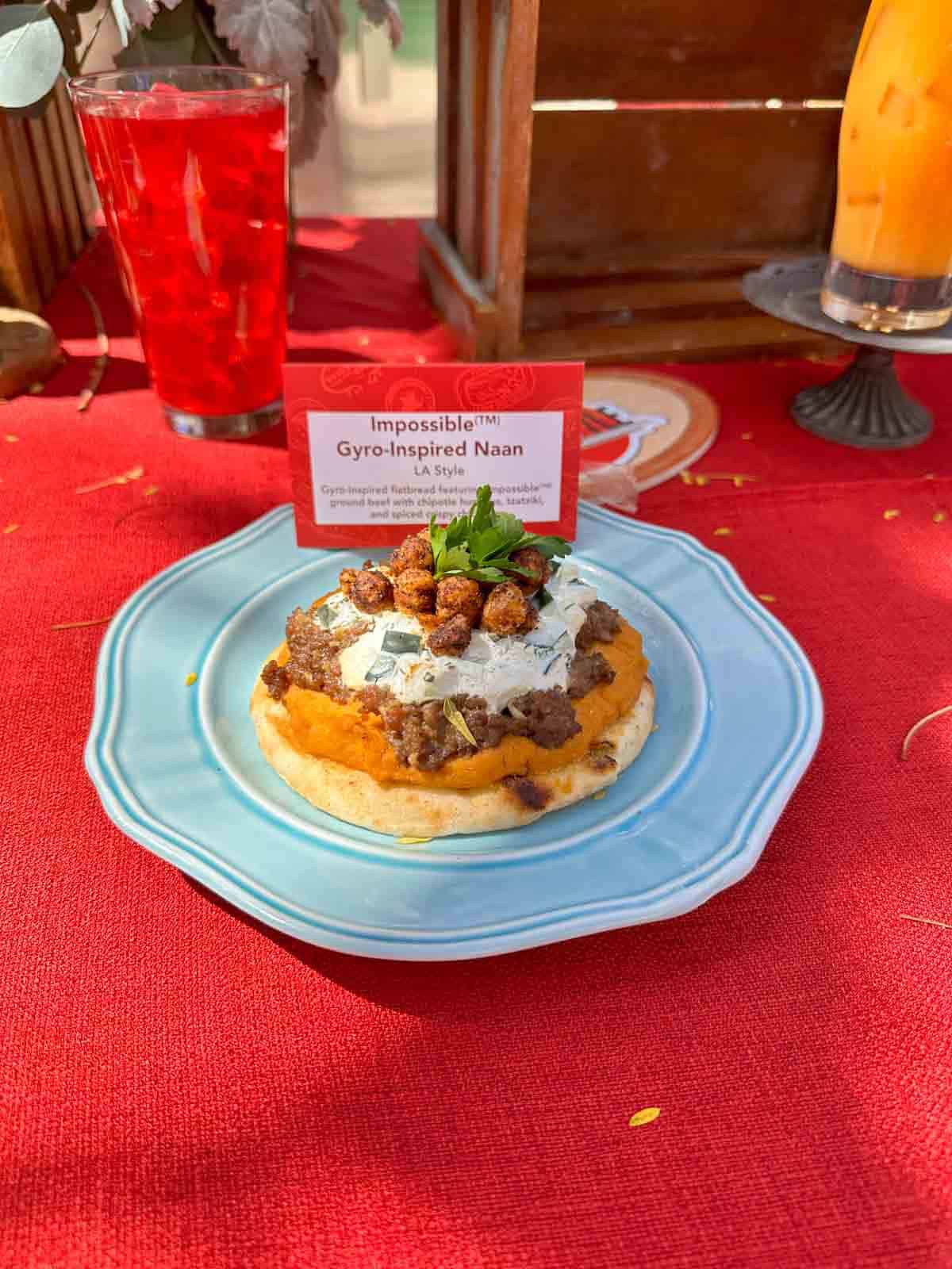 A piece of round naan bread with hummus, ground meat, tzatziki sauce and crispy chickpeas on a blue plate.