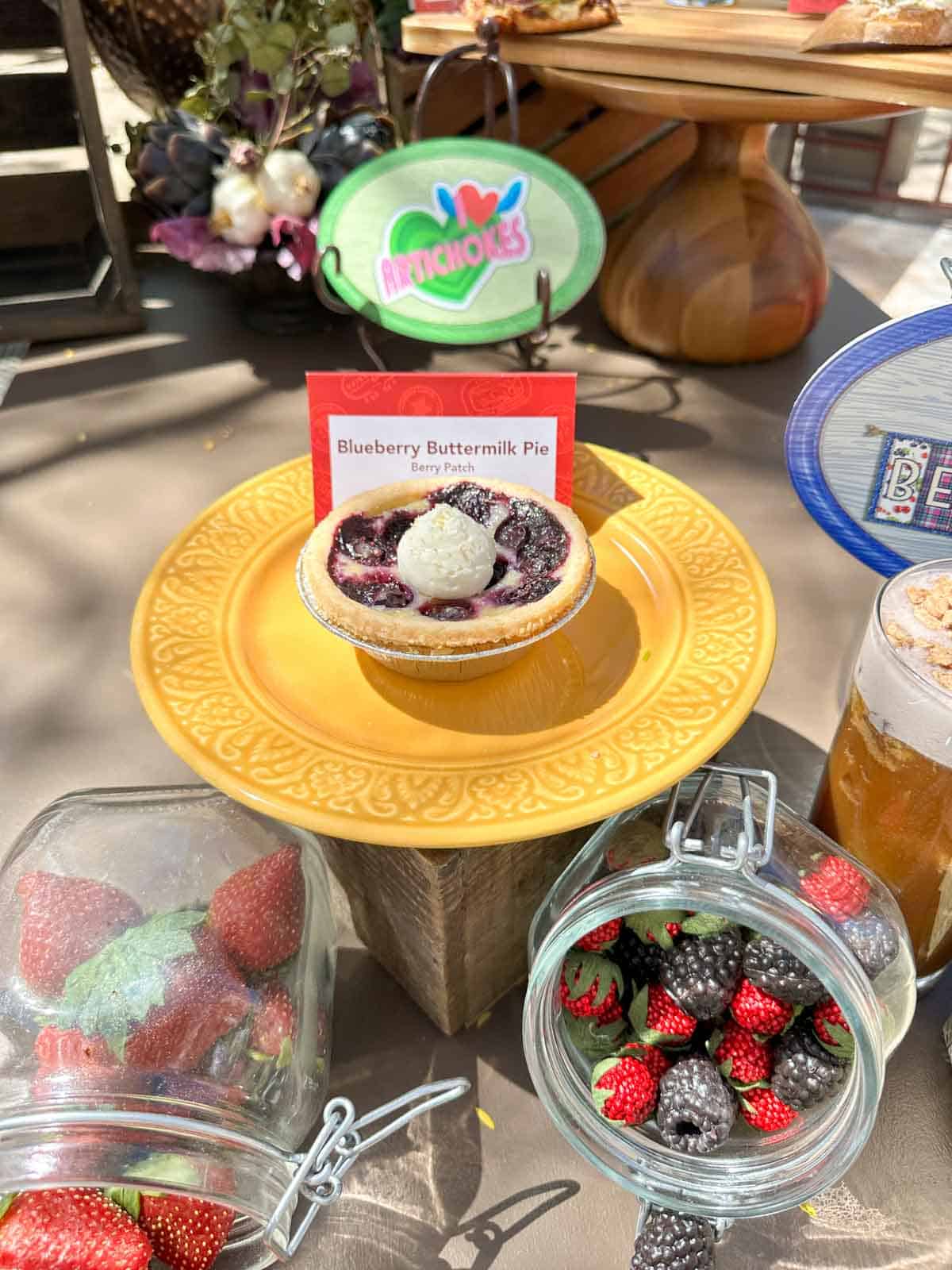 A mini pie with blueberries and cream on a yellow plate sitting on a stand outside on a table.