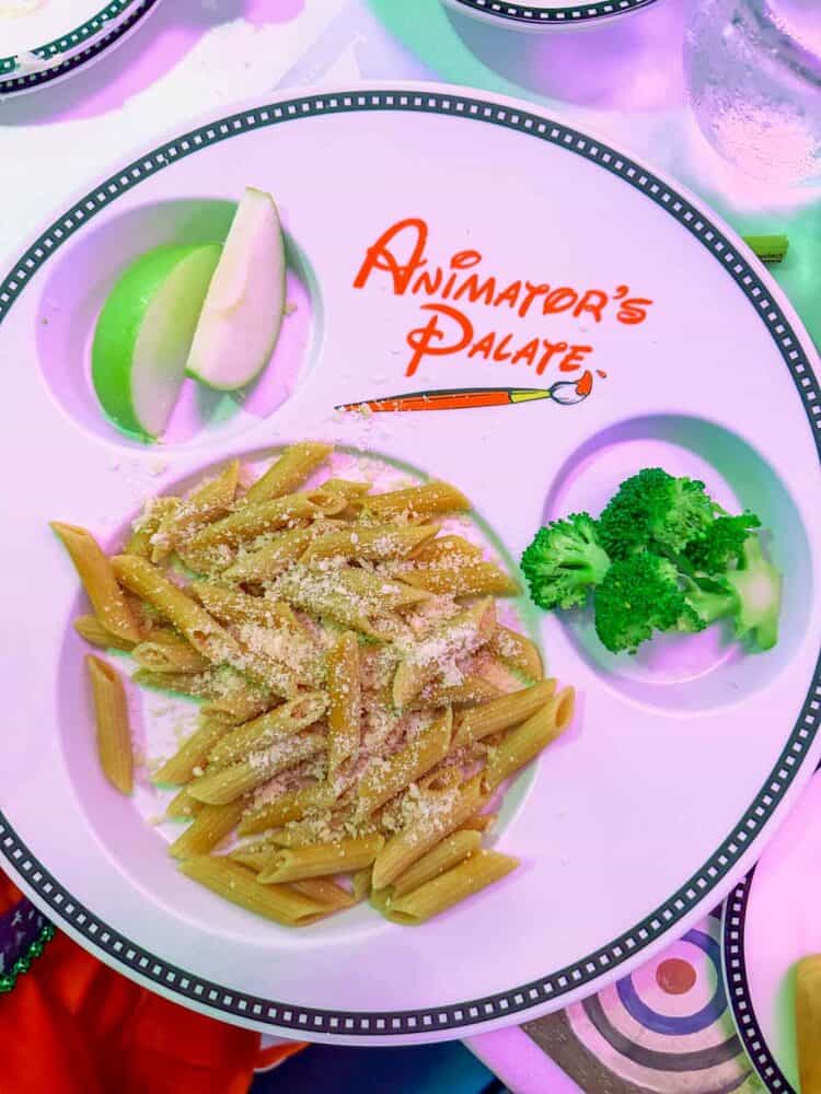 Top view of a white plate with Mickey shaped holes filled with pasta, apple slices and broccoli.