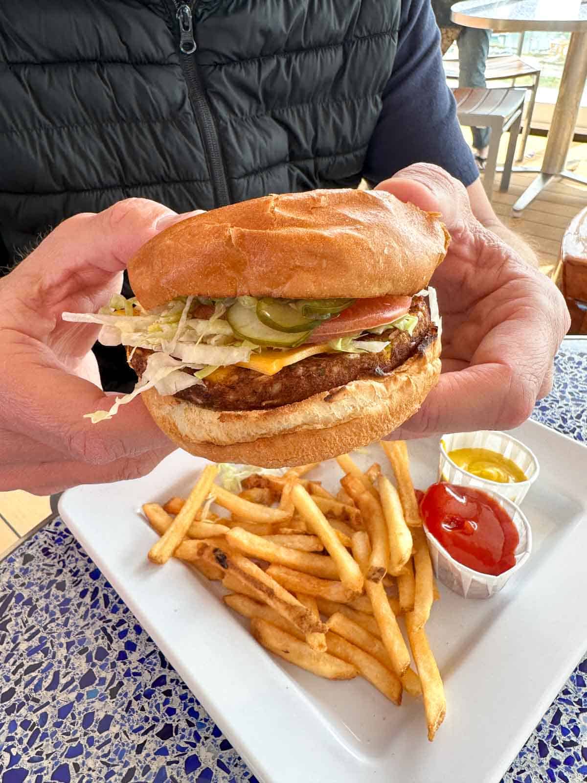 A man holding a burger with toppings on both sides over a white plate of fries.