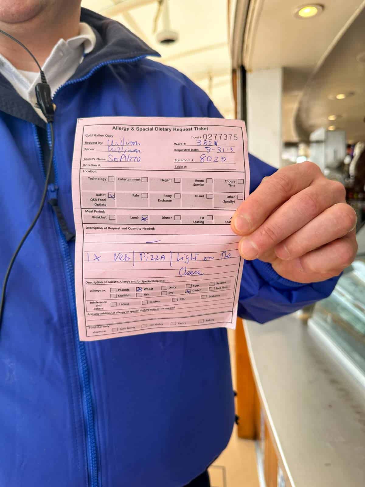 A man in a blue jacket holding up a pink slip of paper with a persons info and dietary accommodations needed.