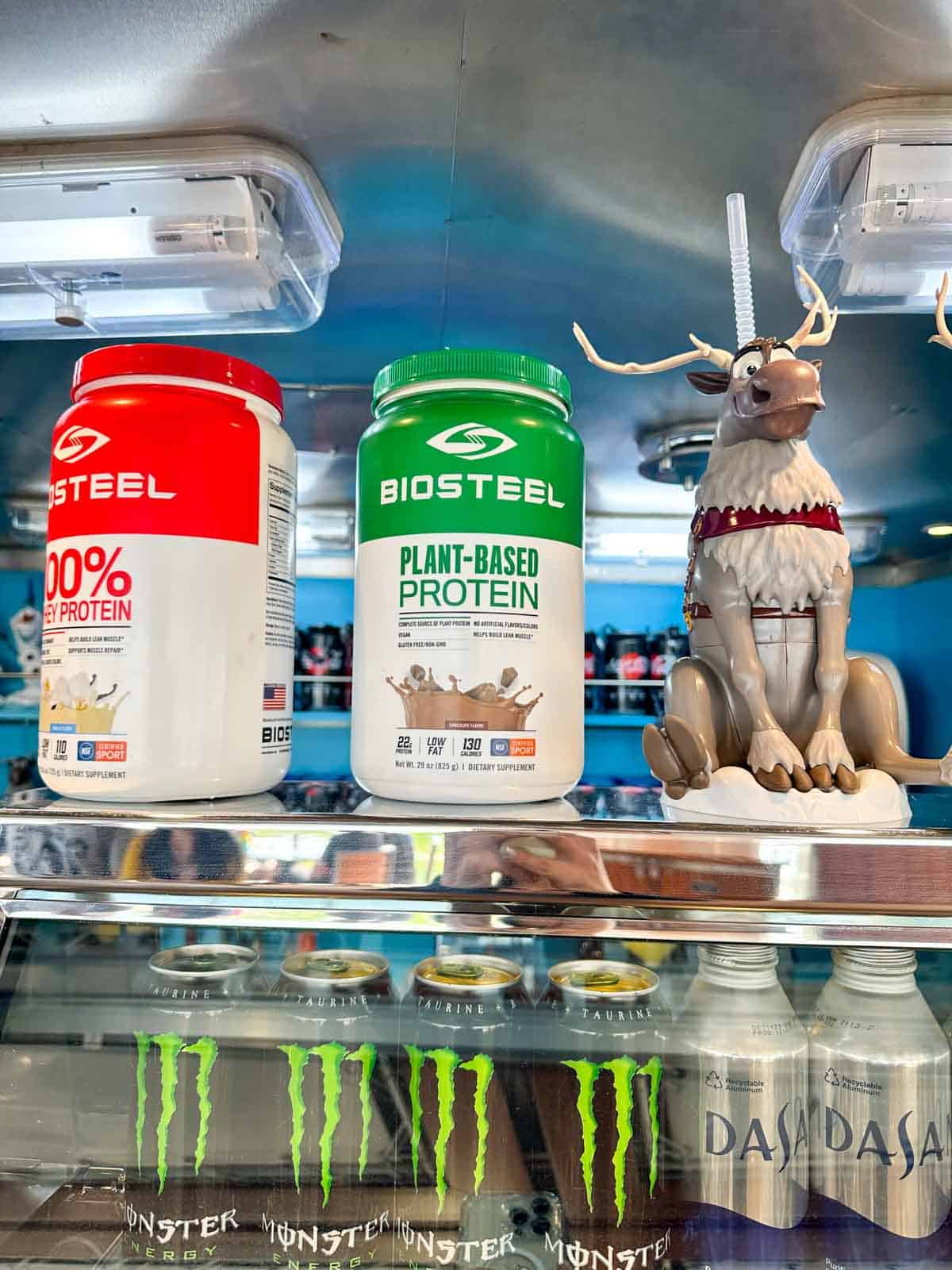 Two containers of protein powder on top of a glass case of canned drinks.