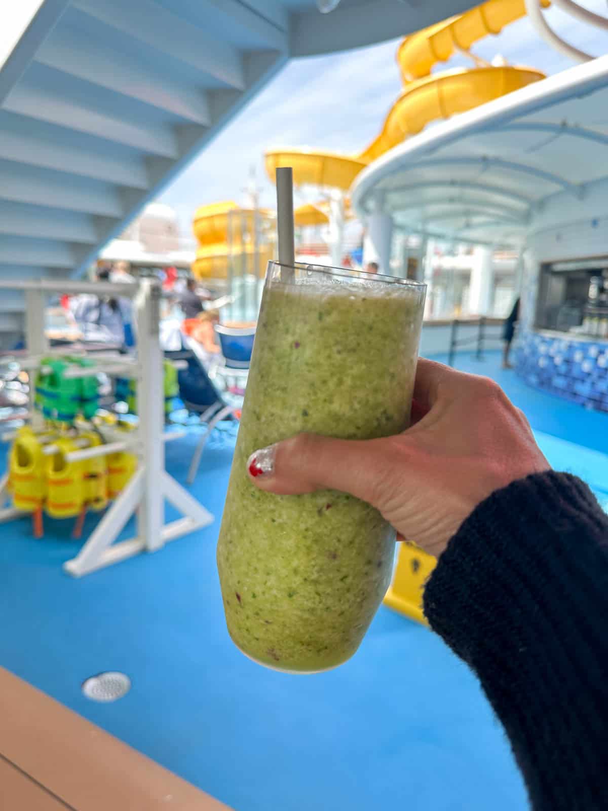 A hand holding a green smoothie in front of a cruise ship pool area.