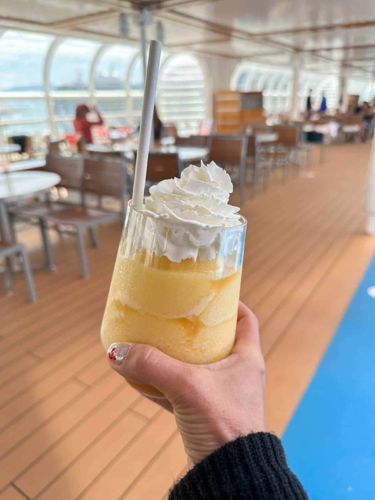 A hand holding a yellow frosted drink with whipped cream on top in front of a cruise ship table area.