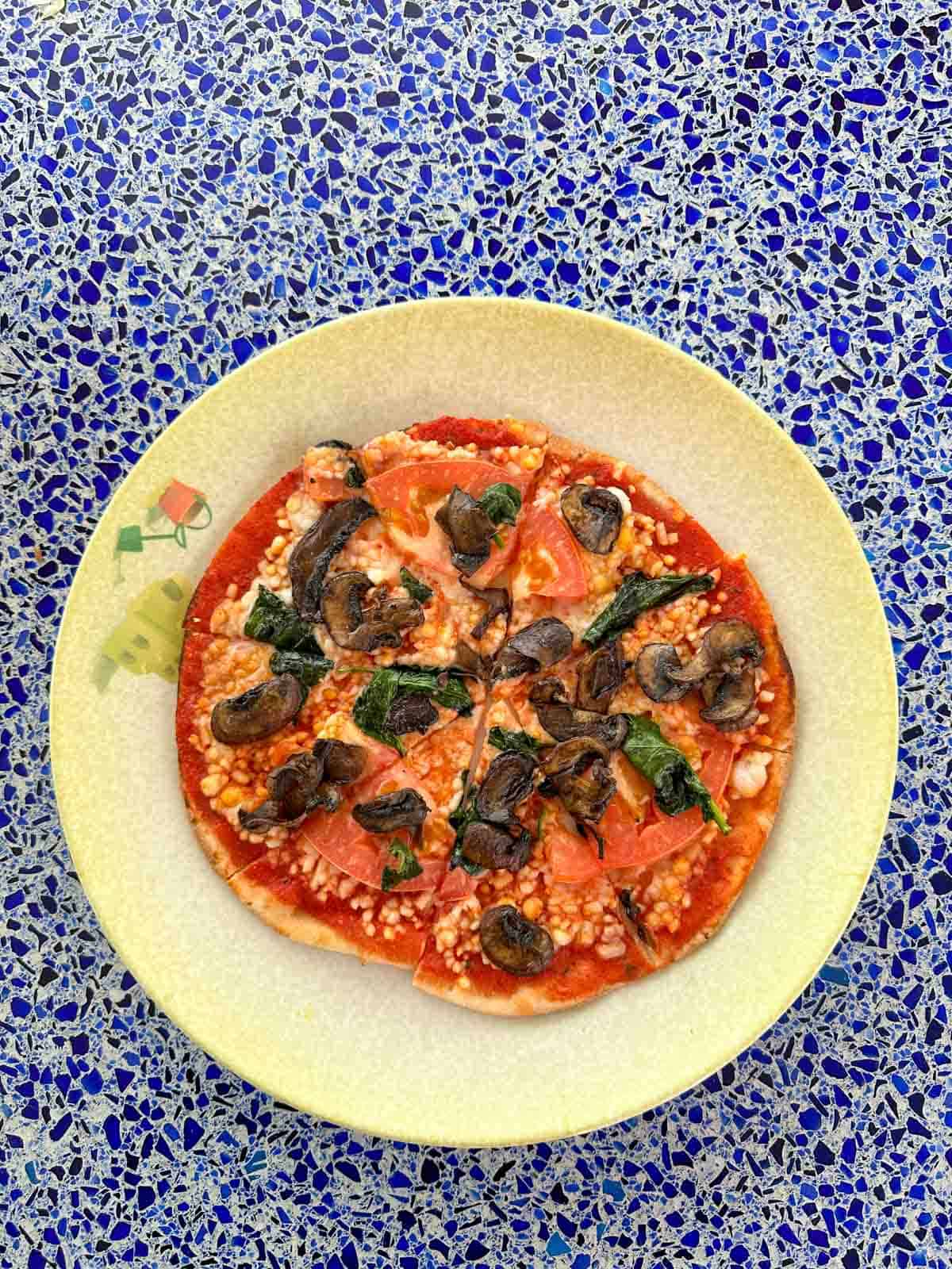 A round pizza with veggies on it on a a yellow plate sitting on a blue speckled table top.