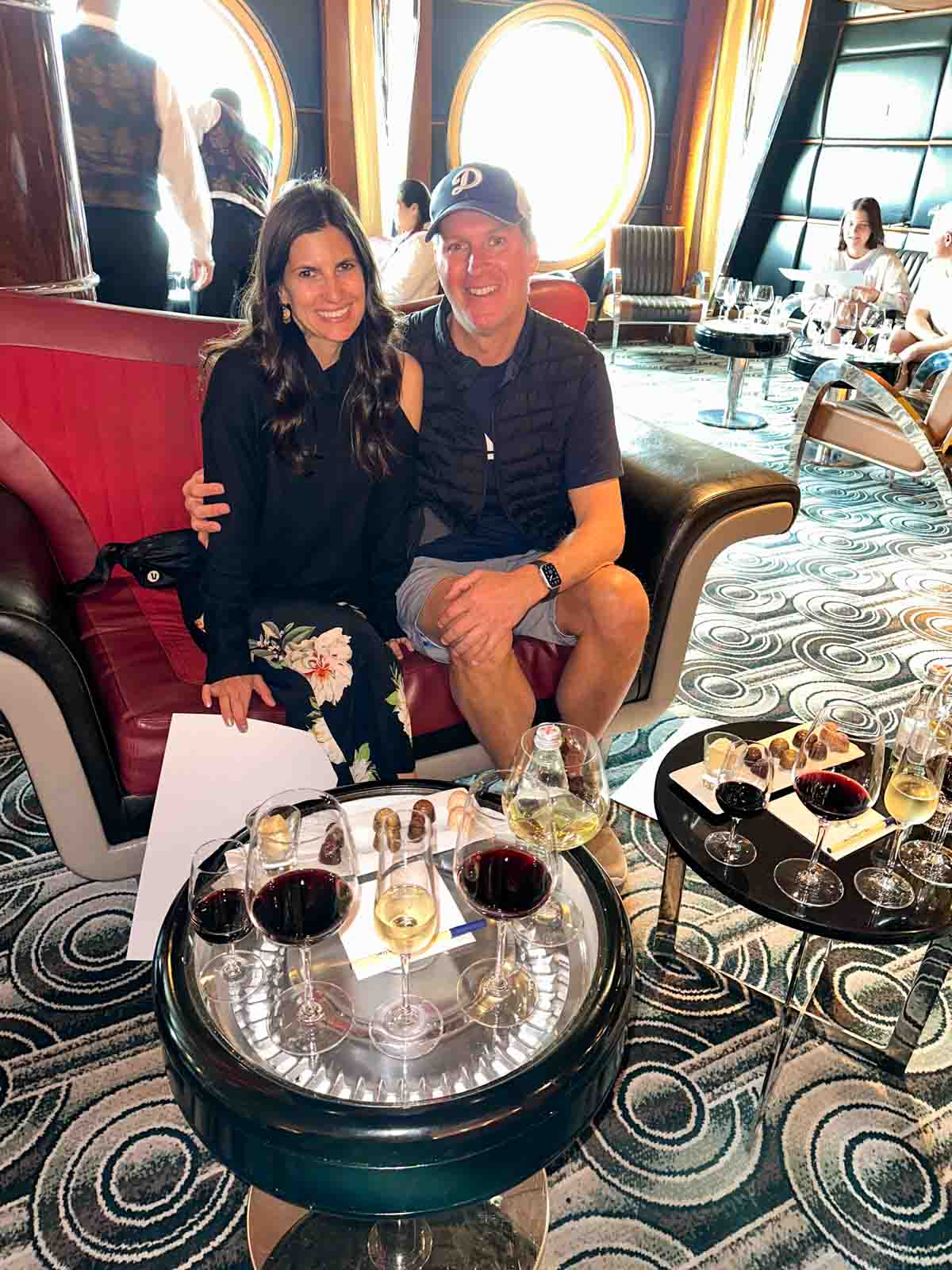 A couple sitting on a red couch in front of two low tables with lined up wine glasses and chocolates on them.