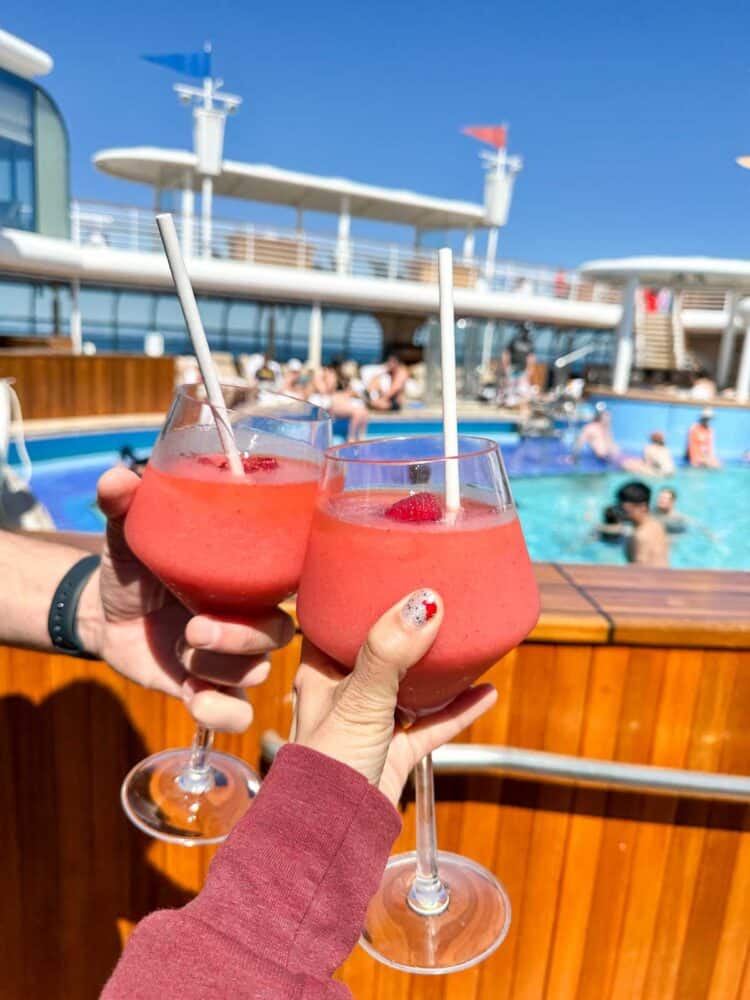 Hands holding glasses with red frosted drinks in front of a pool and saying cheers.