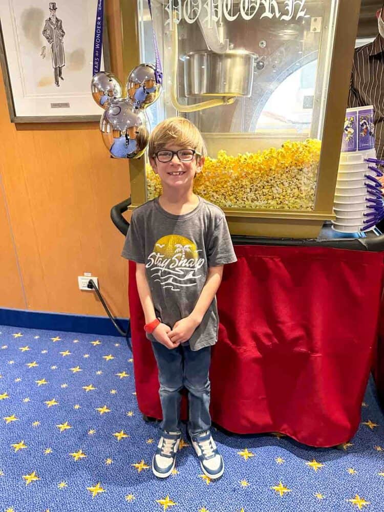 A boy in a grey shirt and jeans in front of a red tablecloth covered table with a popcorn machine on top.