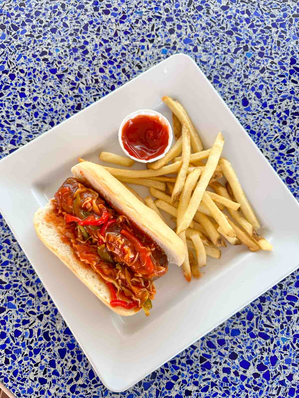 A square white plate with french fries and a sausage dog covered in a red sauce.