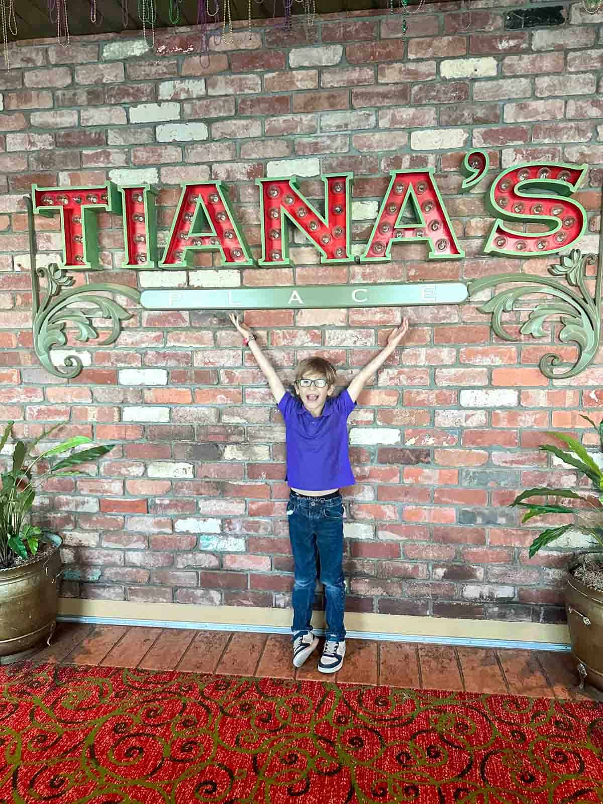 A boy in a purple polo shirt and jeans under a sign saying Tiana's on a bring wall.