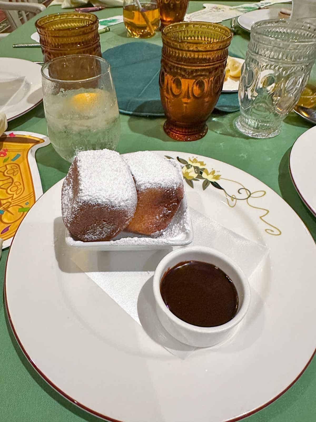 A white plate with a bowl of chocolate sauce and a bowl of two powdered sugar covered beignets.