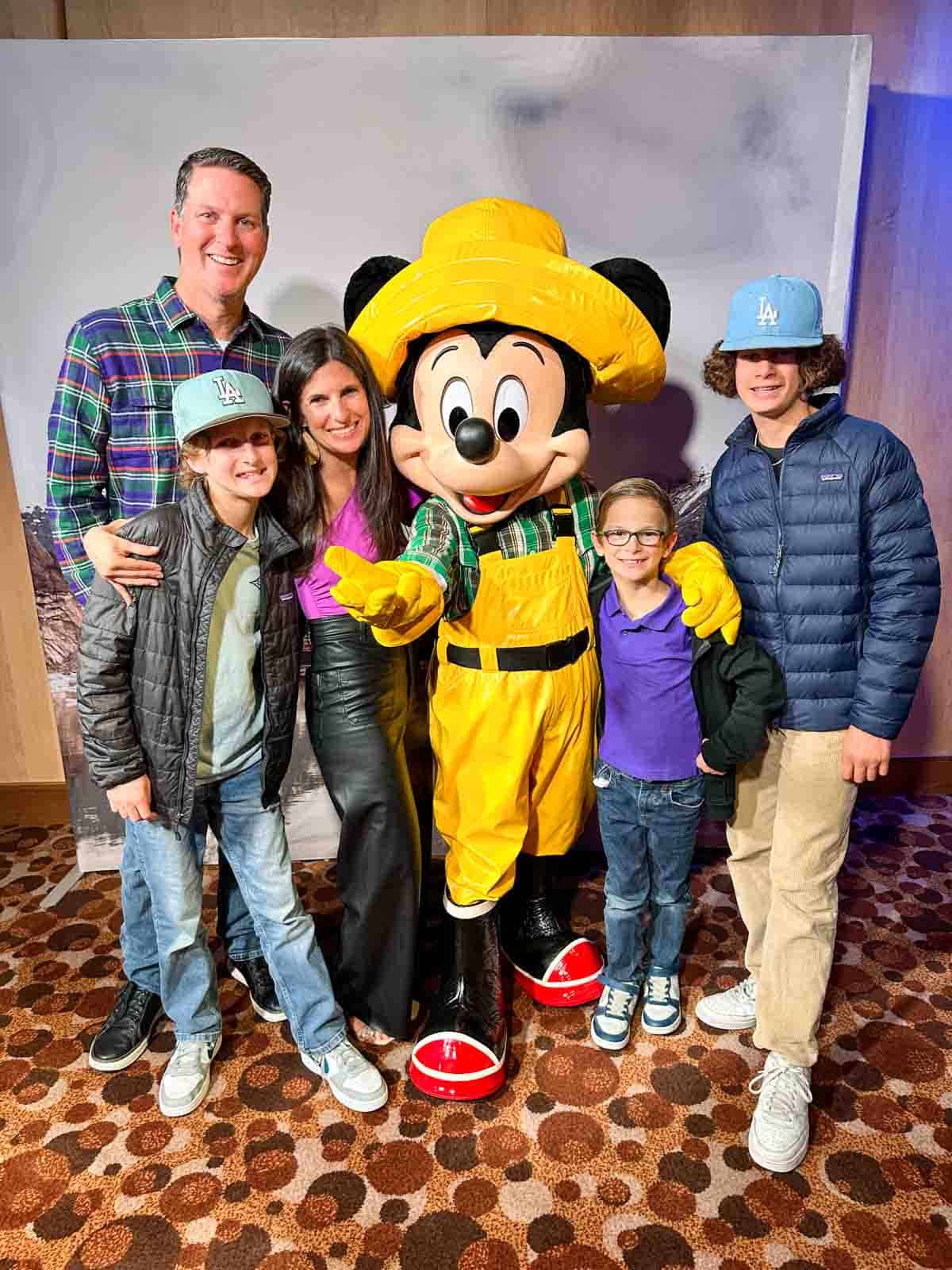 A family of five standing in a room around a live Mickey Mouse character dressed in a yellow hat and fisherman pants taking a photo.