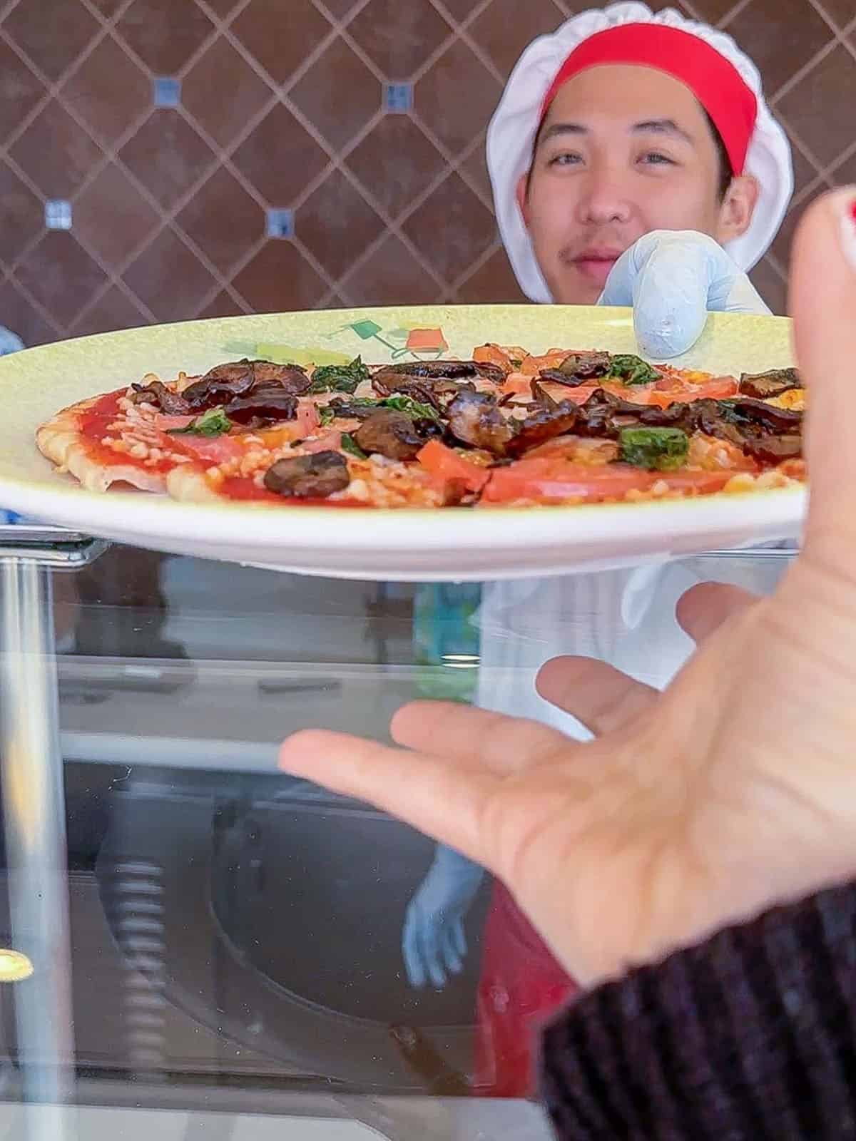 A chef handing someone a pizza on a yellow plate over a glass covered counter.