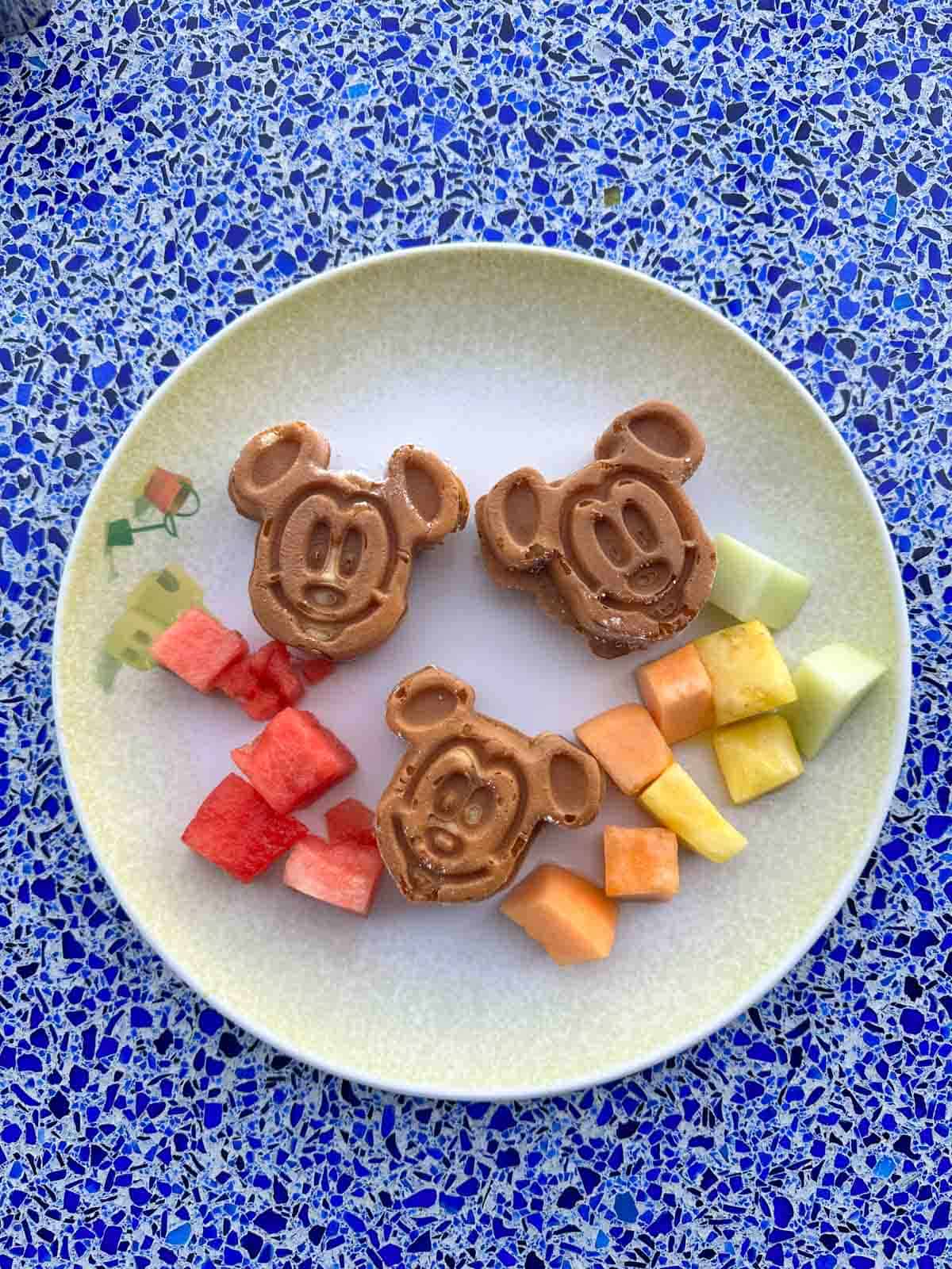 Top view of Mickey Mouse shaped waffles on white plate with fruit on it too.