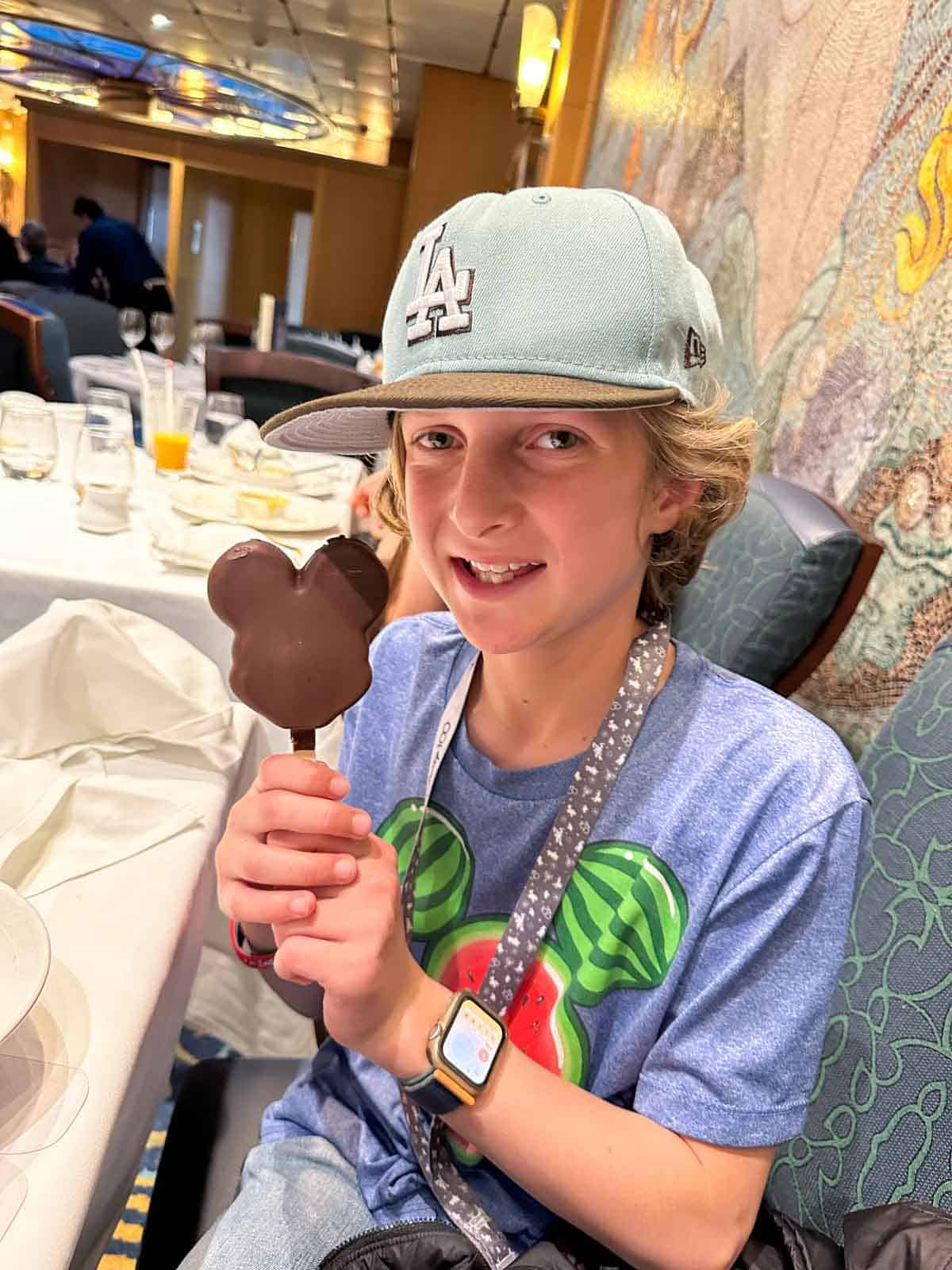 A boy in a light green blue hat holding a chocolate covered mickey shaped ice cream popsicle.