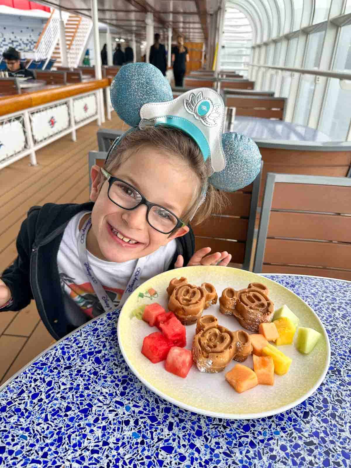 A boy wearing blue Mickey ears and glasses smiling big in front of a plate with mini Mickey waffles and fruit on it.
