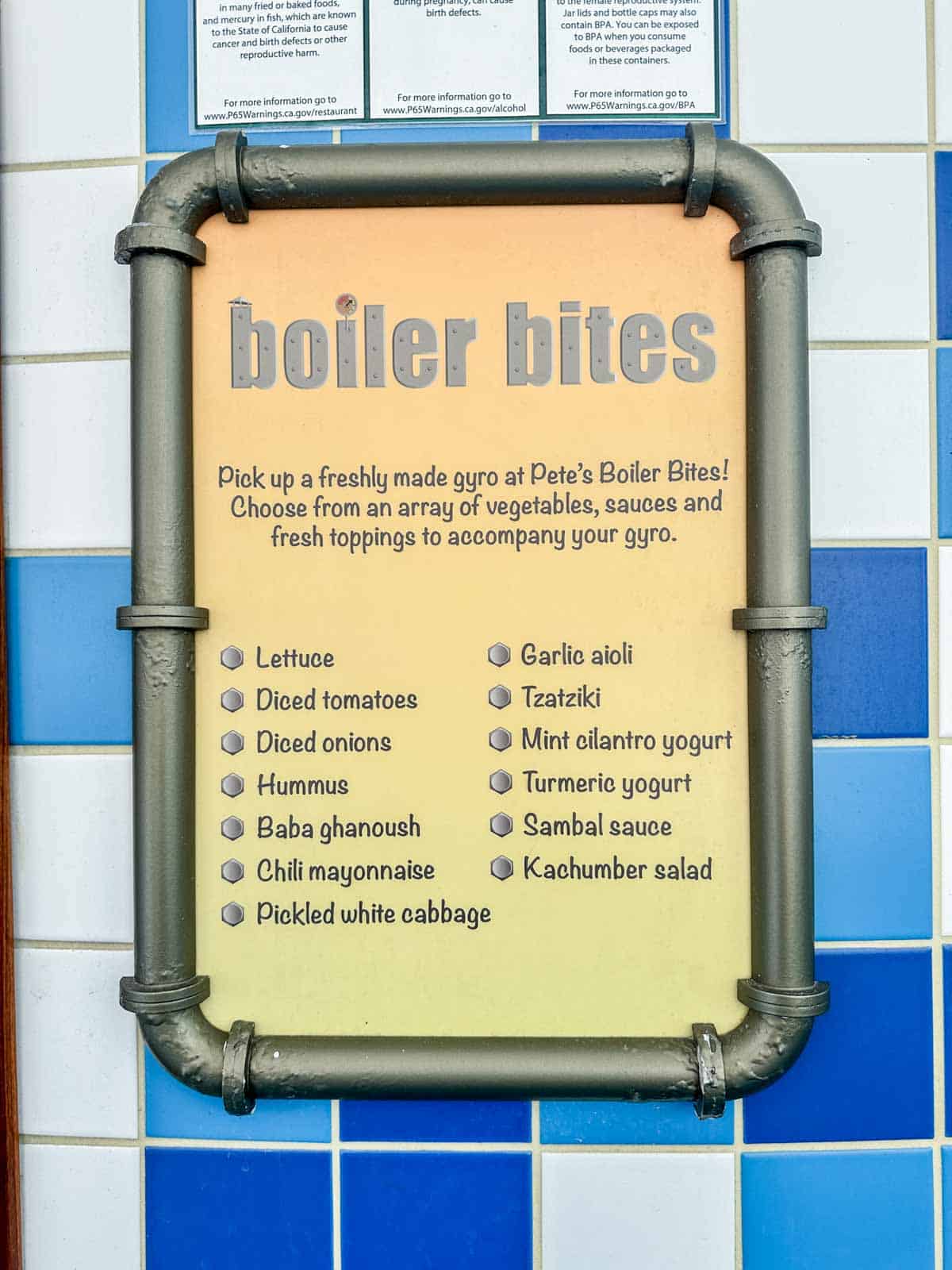 A yellow sign with pipes framed around with grey writing saying Boiler Bites and a list of items under.