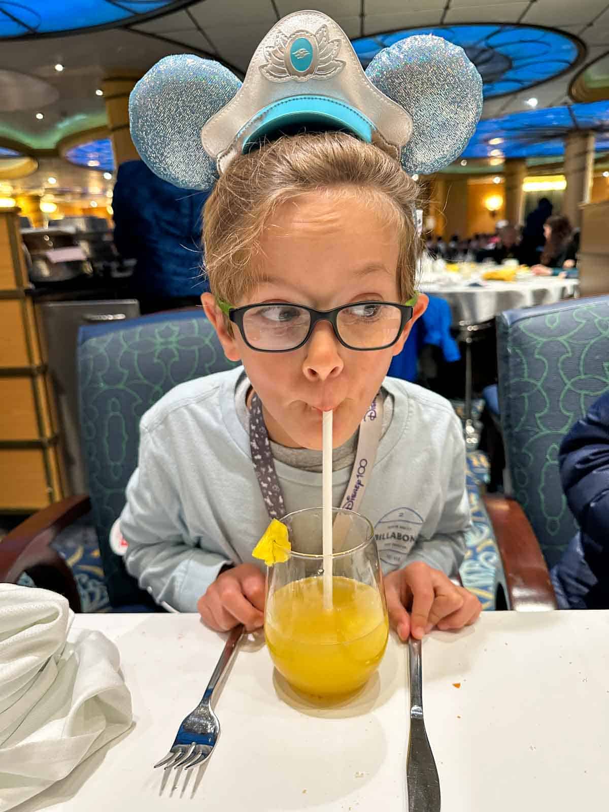 A boy with blue Mickey ears taking a drink from a straw in a glass with yellow juice in it.