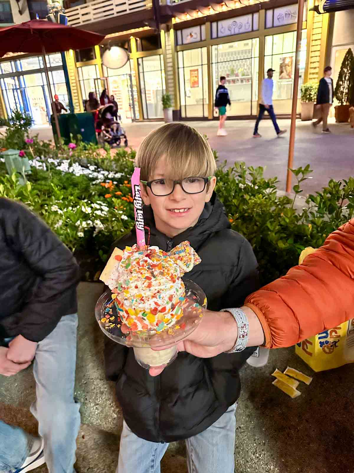 A hand holding a sundae like shake topped with colorful toppings in front of a little blond boy at night.