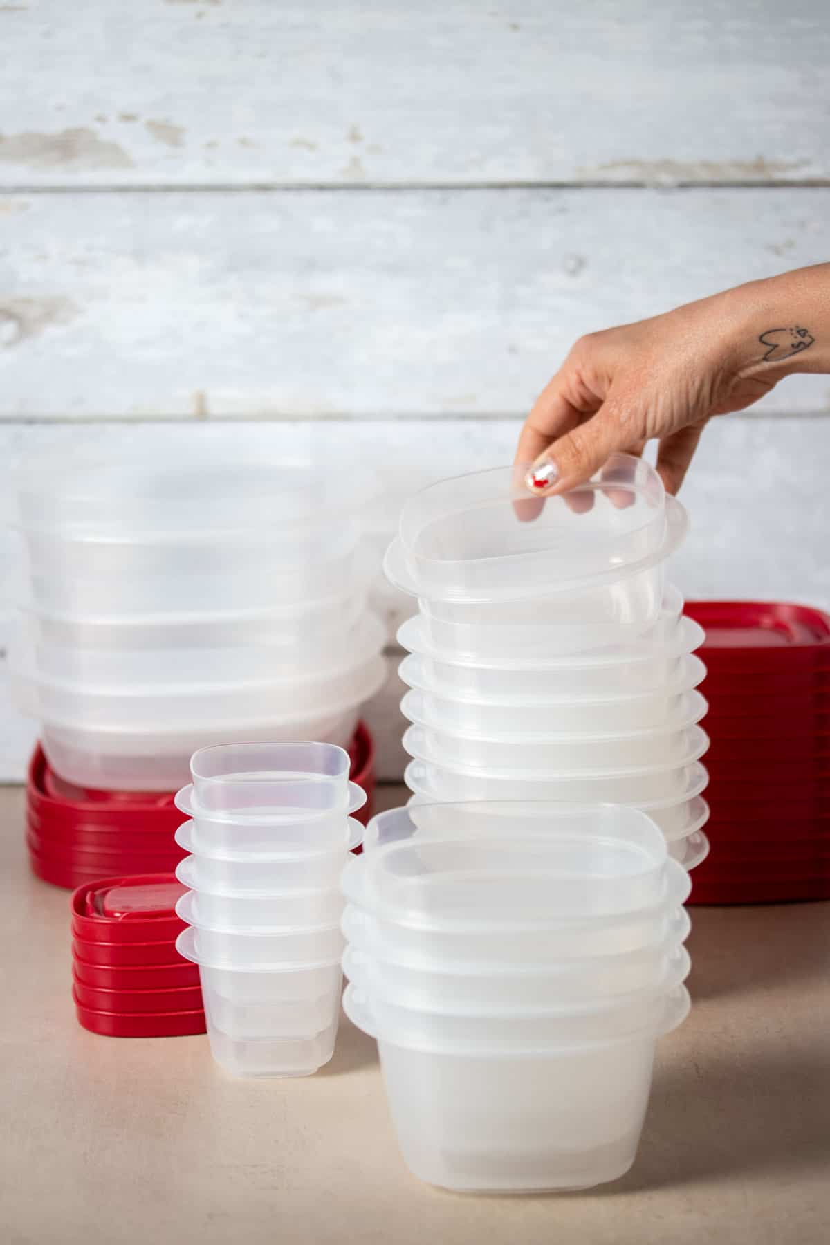 A hand grabbing a plastic container from a stack of them with red lids on a tan surface.