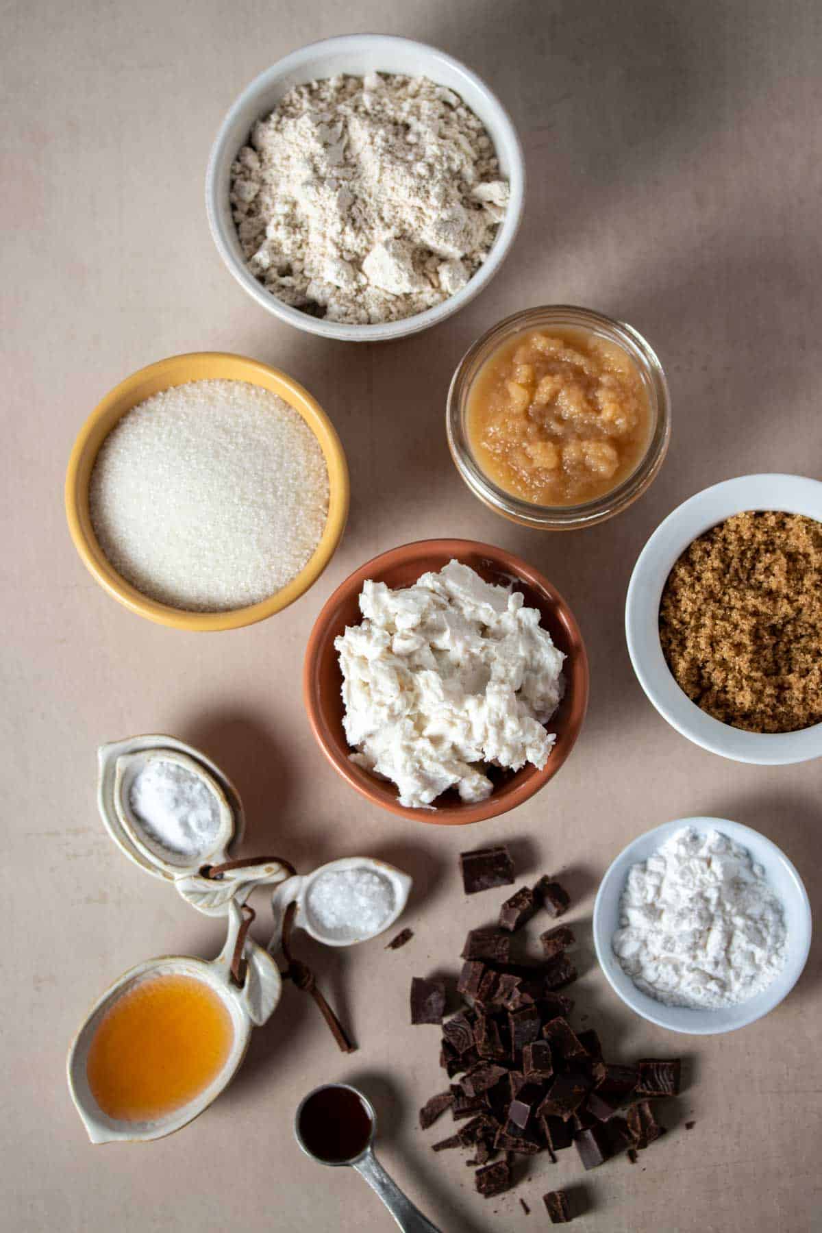 Top view of different bowls with ingredients to make chocolate chip cookies on a tan surface.