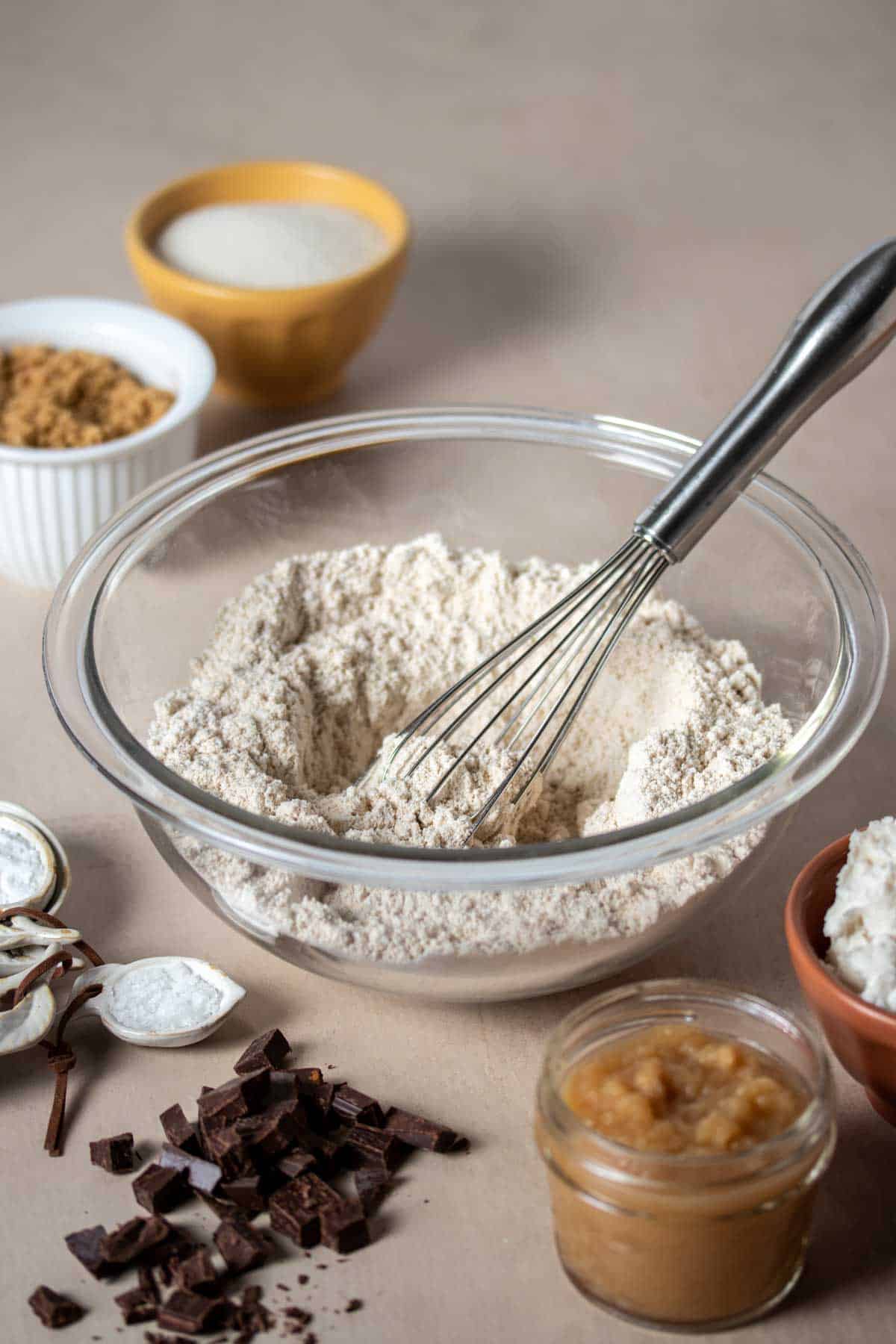 A glass bowl with flour in it being mixed with a which sitting on a tan surface with ingredients around it.