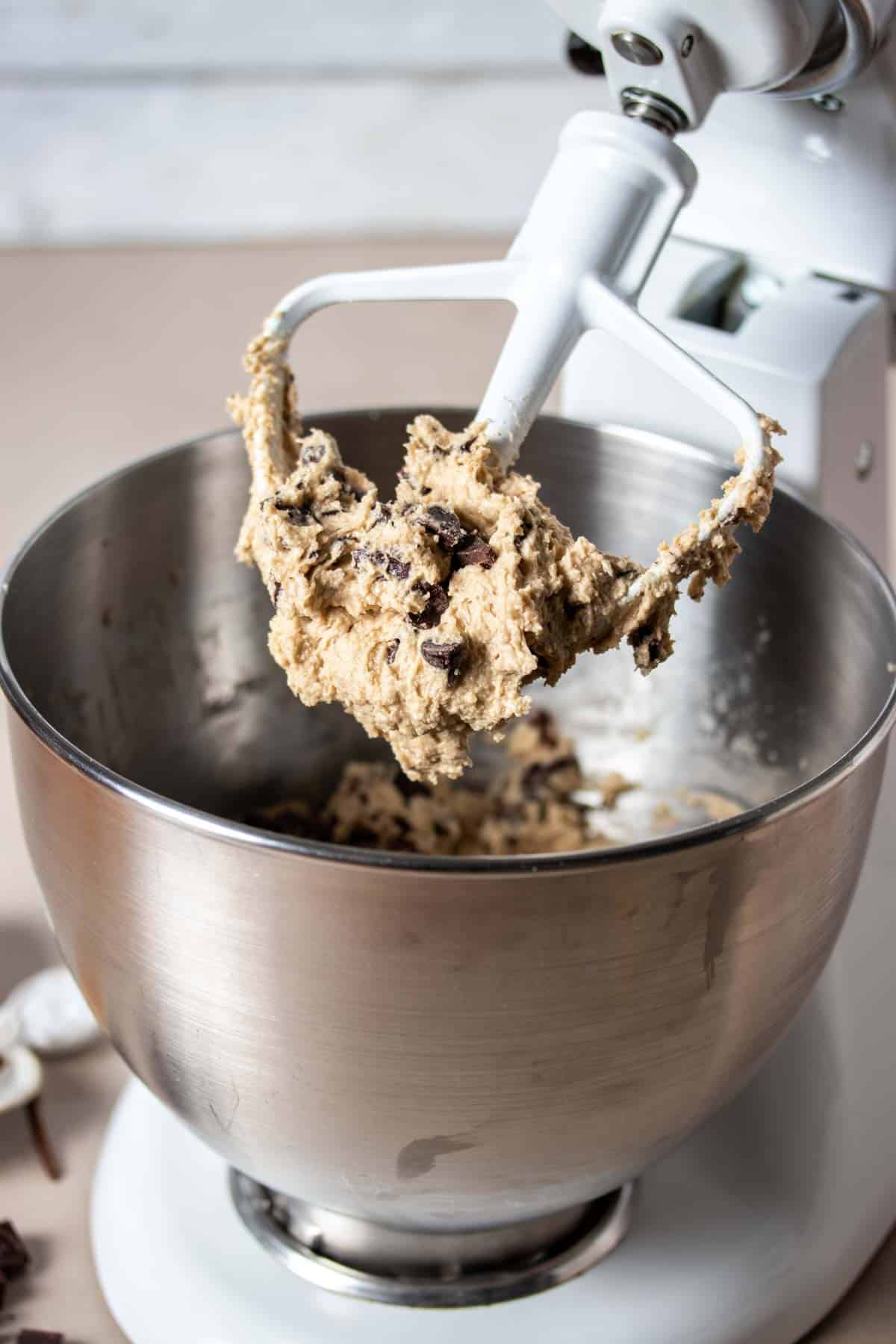 A white mixer paddle with chocolate chip cookie dough on it coming out of a silver mixer bowl.