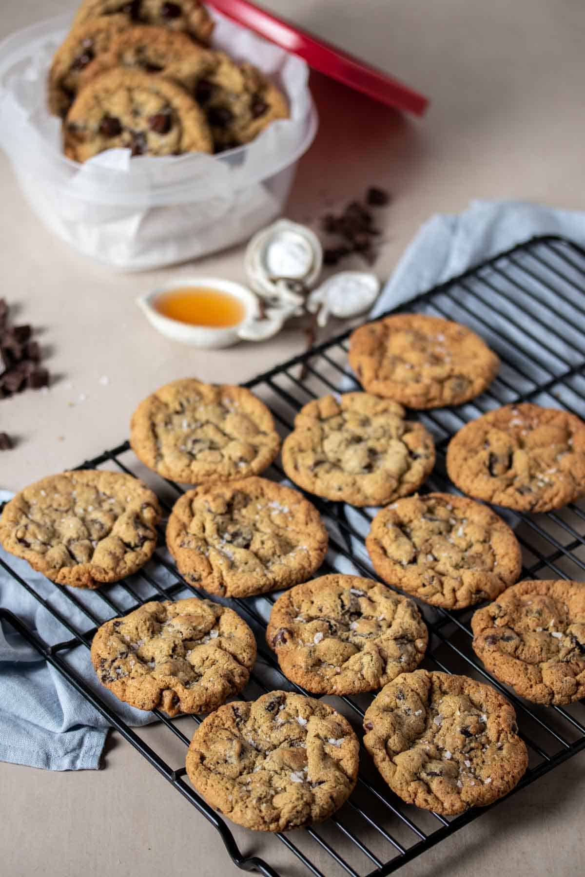 A black wire cooling rack lined with chocolate chip cookies on a blue towel with a container of cookies in the back.