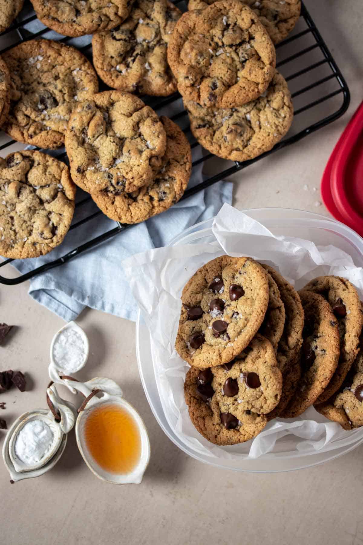 Top view of chocolate chip cookies in a parchment lined plastic container on a tan surface next to a rack of cookies.